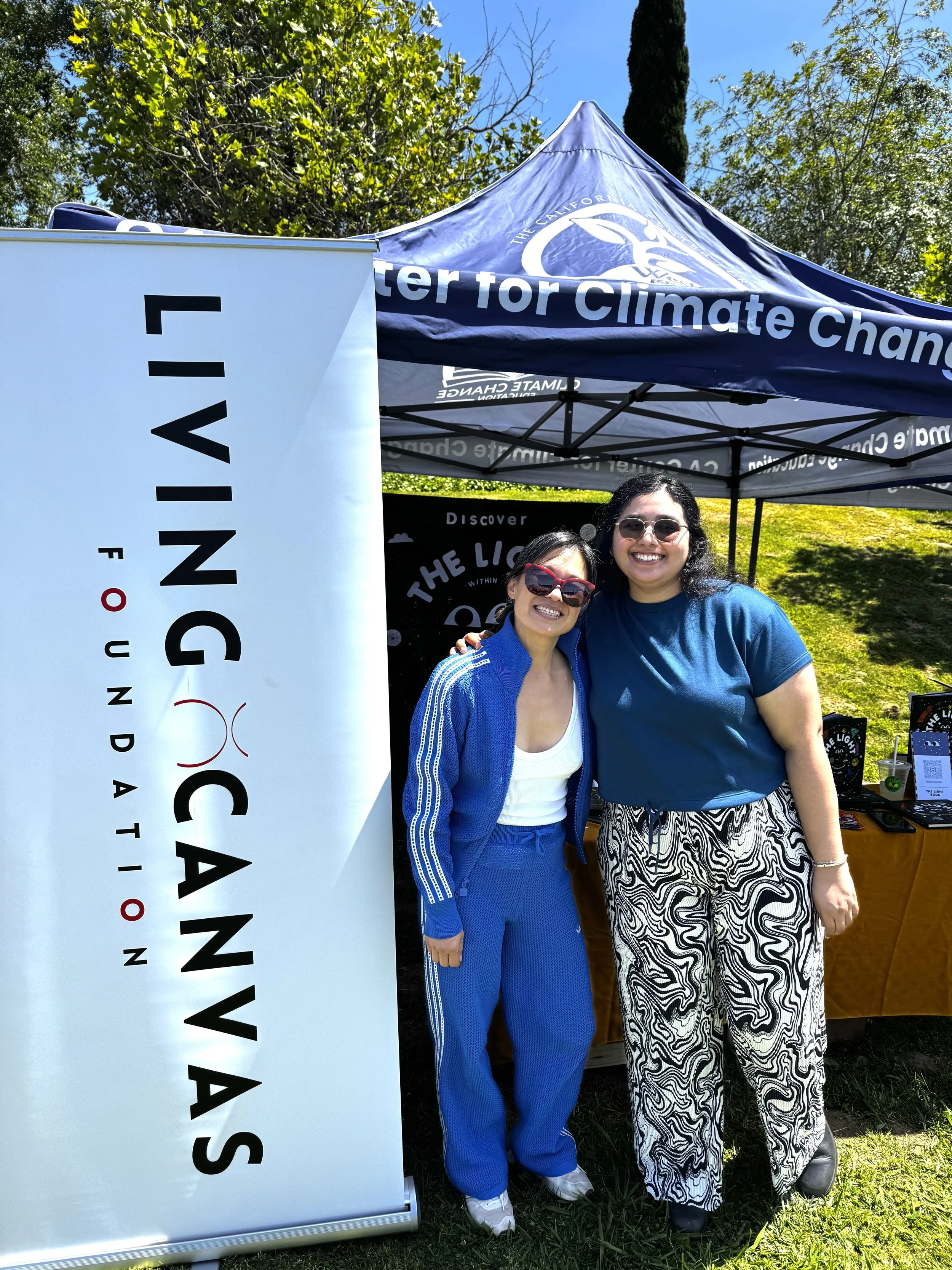 Two women smiling and standing outside under a blue canopy tent that has a logo and text about climate change. One woman is wearing a blue tracksuit and sunglasses, and the other is wearing a blue T-shirt with black and white patterned pants and sunglasses. There is a large white vertical sign with black and red text partially visible on the left side of the image. The background includes green trees, a grass lawn, and a clear blue sky.