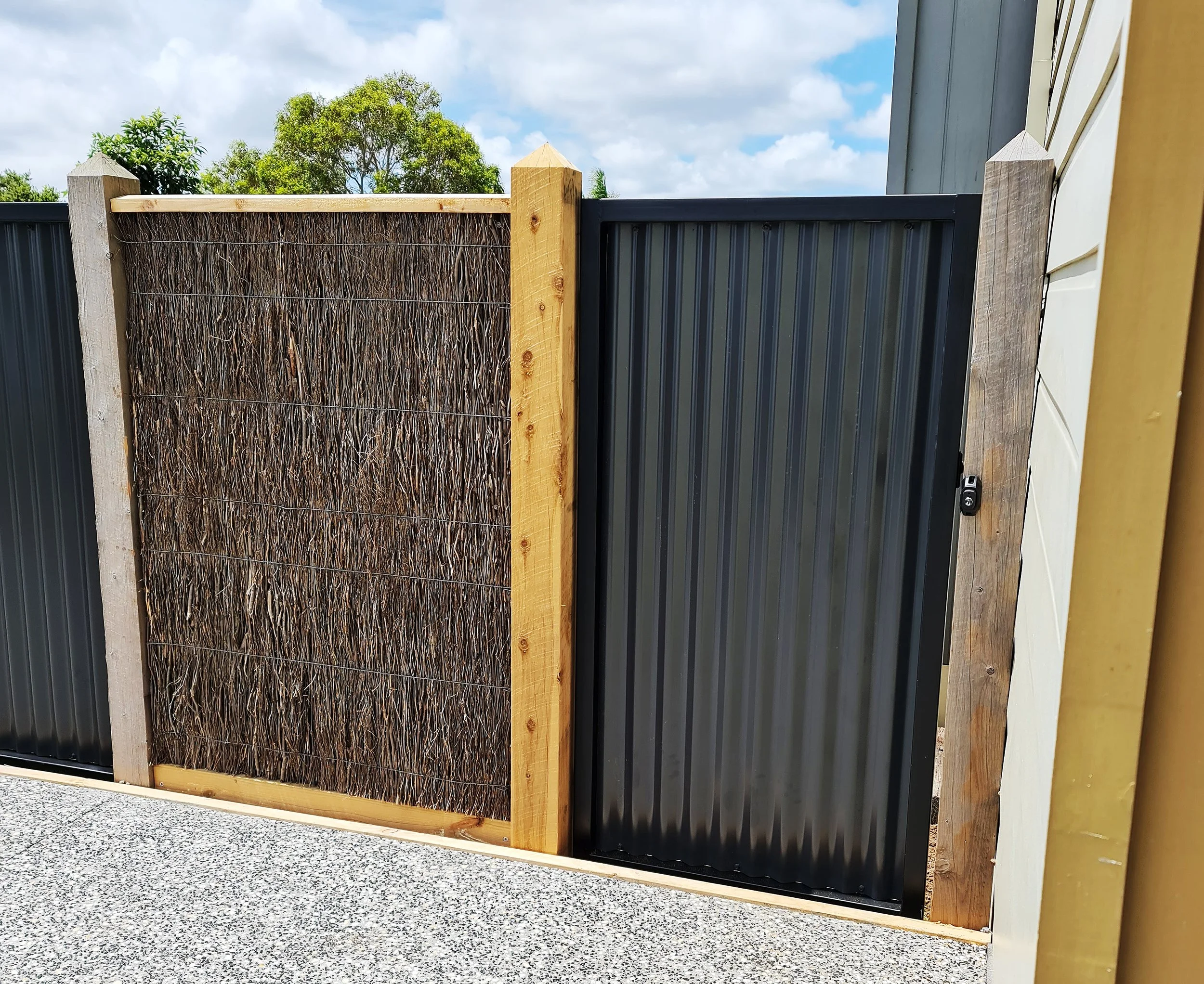 A black corrugated metal gate with a latch, adjacent to a wooden fence section with a decorative woven panel.