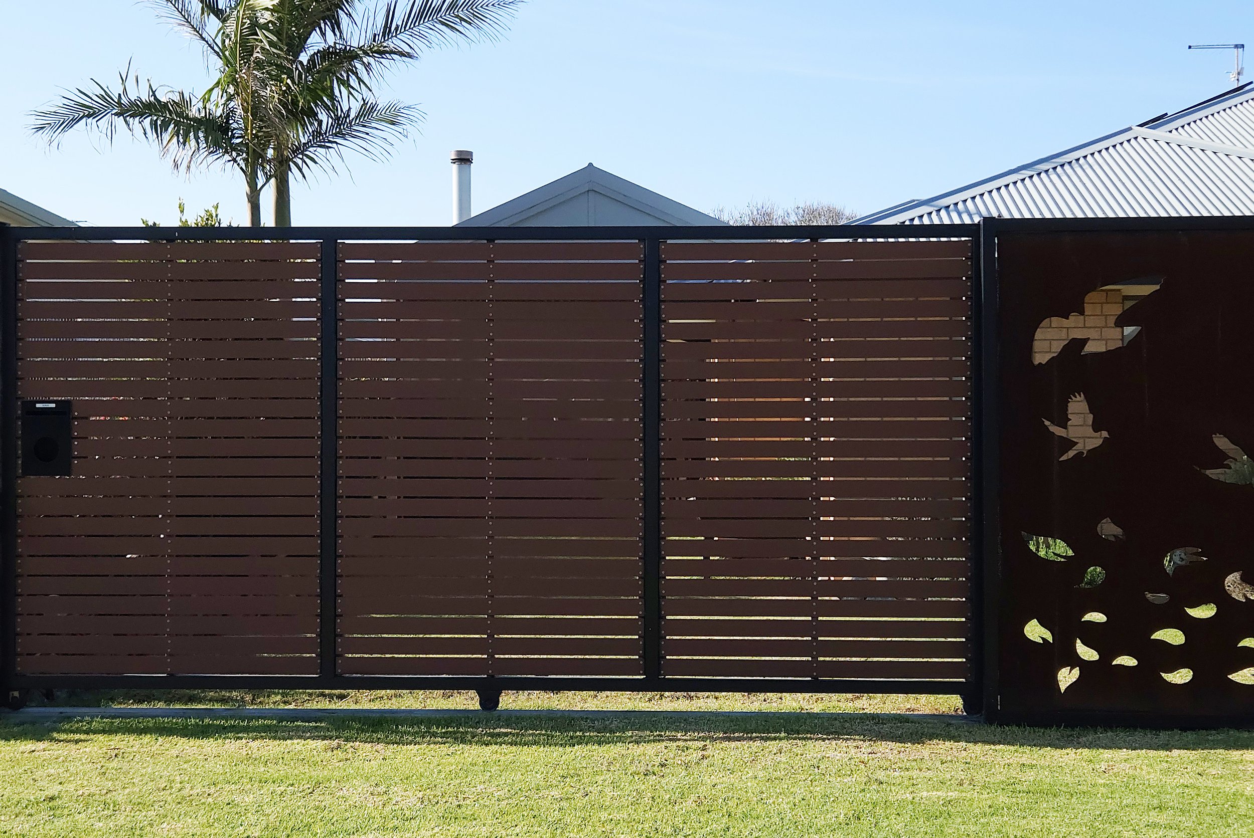 A modern wooden gate with horizontal slats and a decorative patterned panel on the right side, located in a residential yard with green grass and neighboring houses with metal roofs visible in the background.