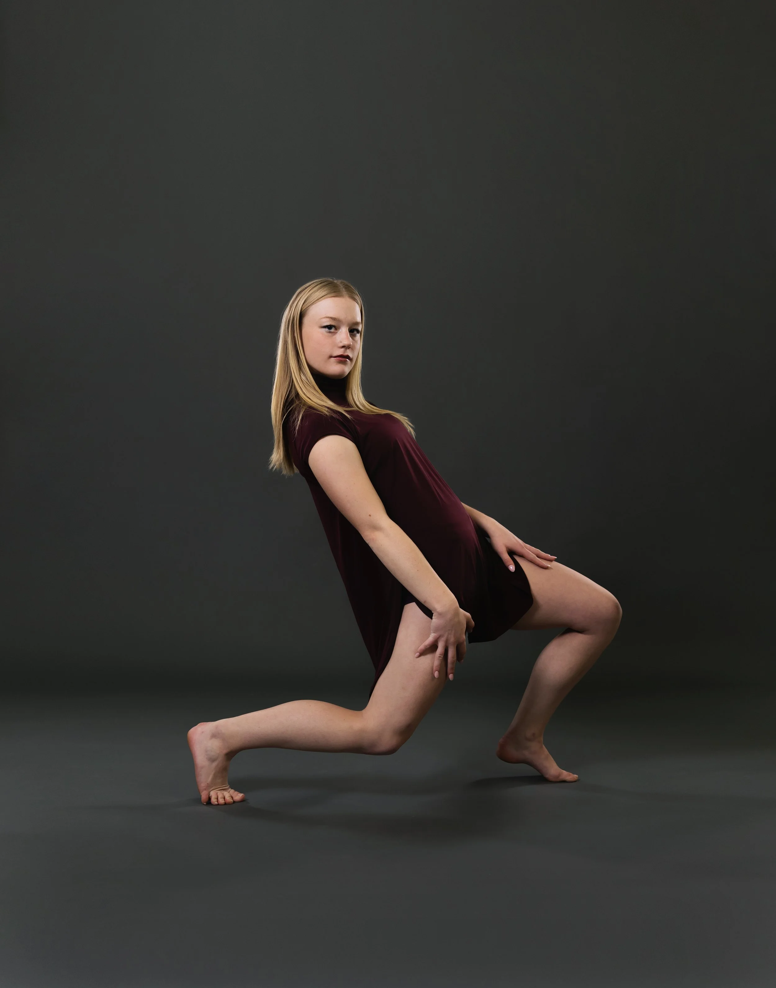 A woman with long blonde hair wearing a burgundy dress practicing yoga on a dark gray background, in a lunge pose with one knee bent and one leg extended behind her.