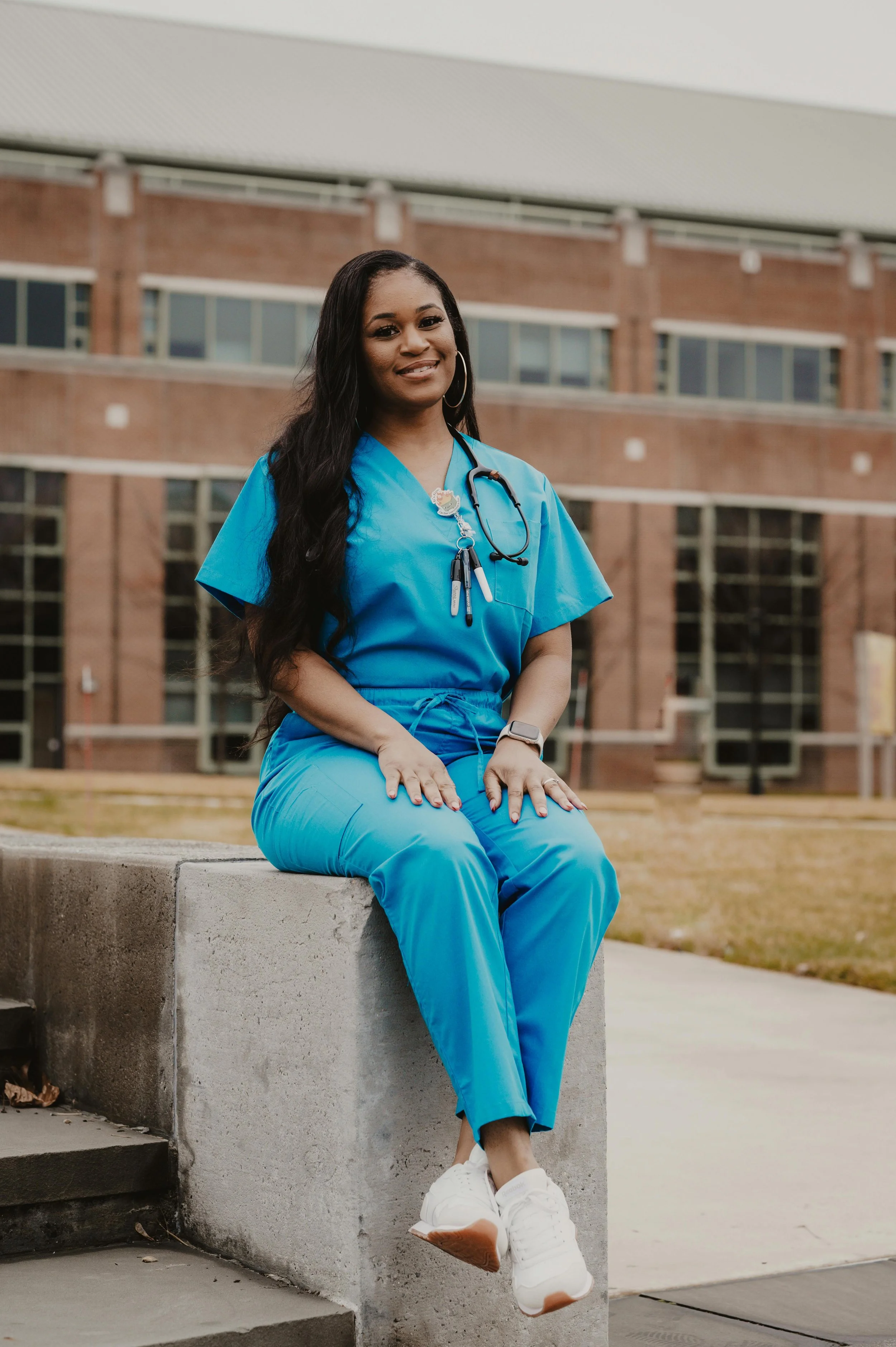 A smiling female nurse in blue scrubs sitting on a concrete ledge outdoors in front of a brick hospital or medical building.