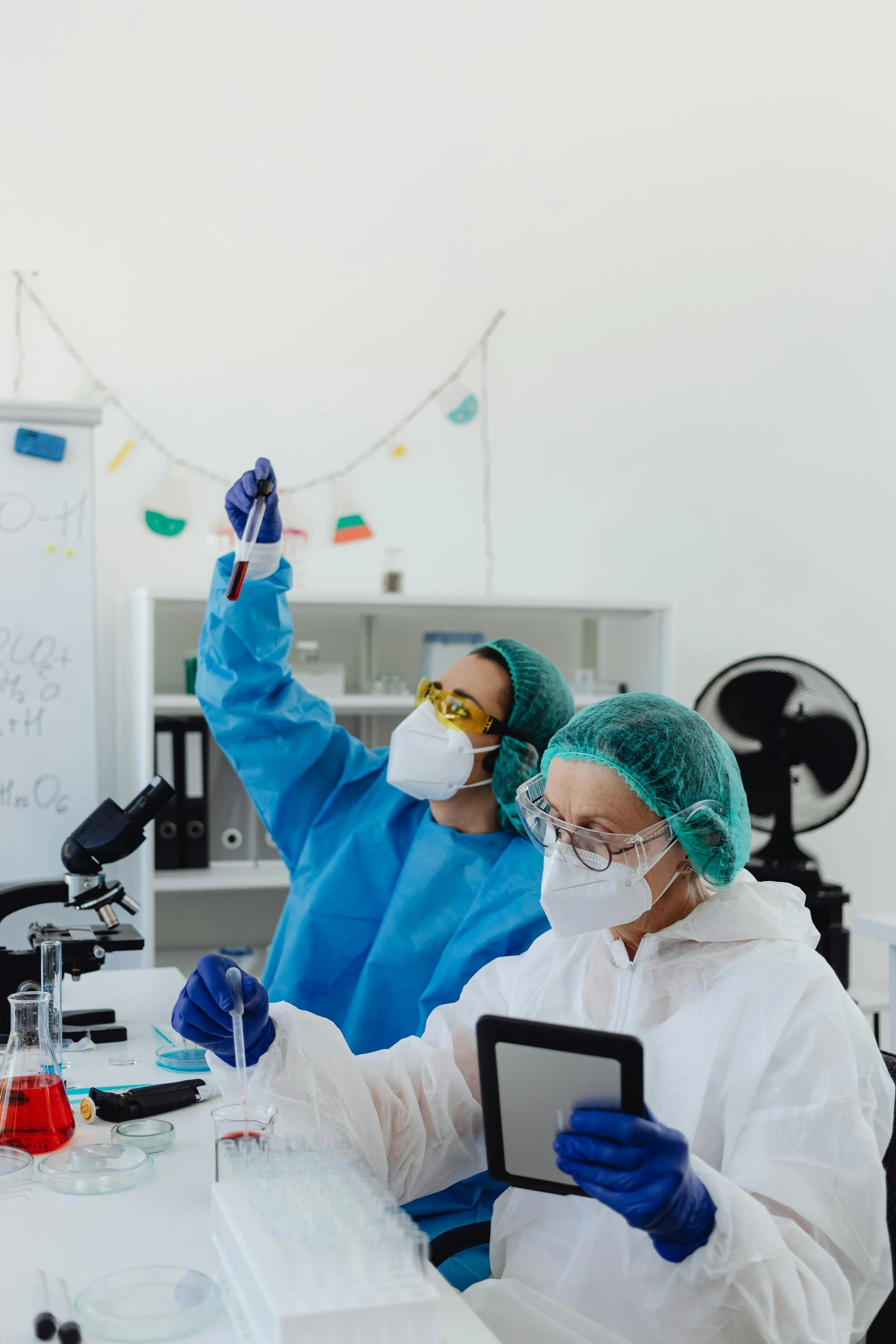 Two scientists wearing protective gear working in a laboratory, one holding a test tube and the other using a tablet.