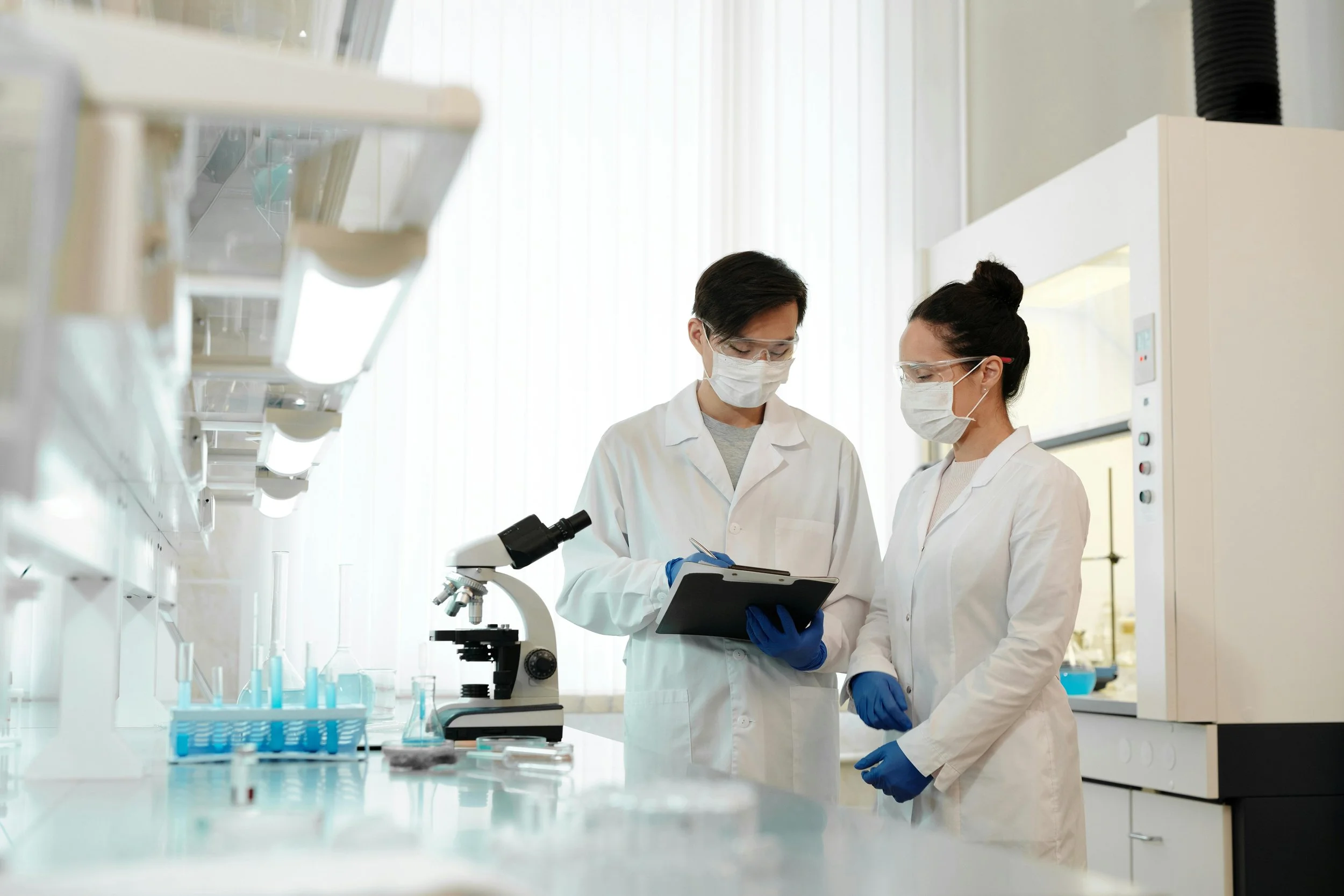 Two scientists wearing masks and safety glasses working in a laboratory, looking at a clipboard near a microscope and laboratory equipment.