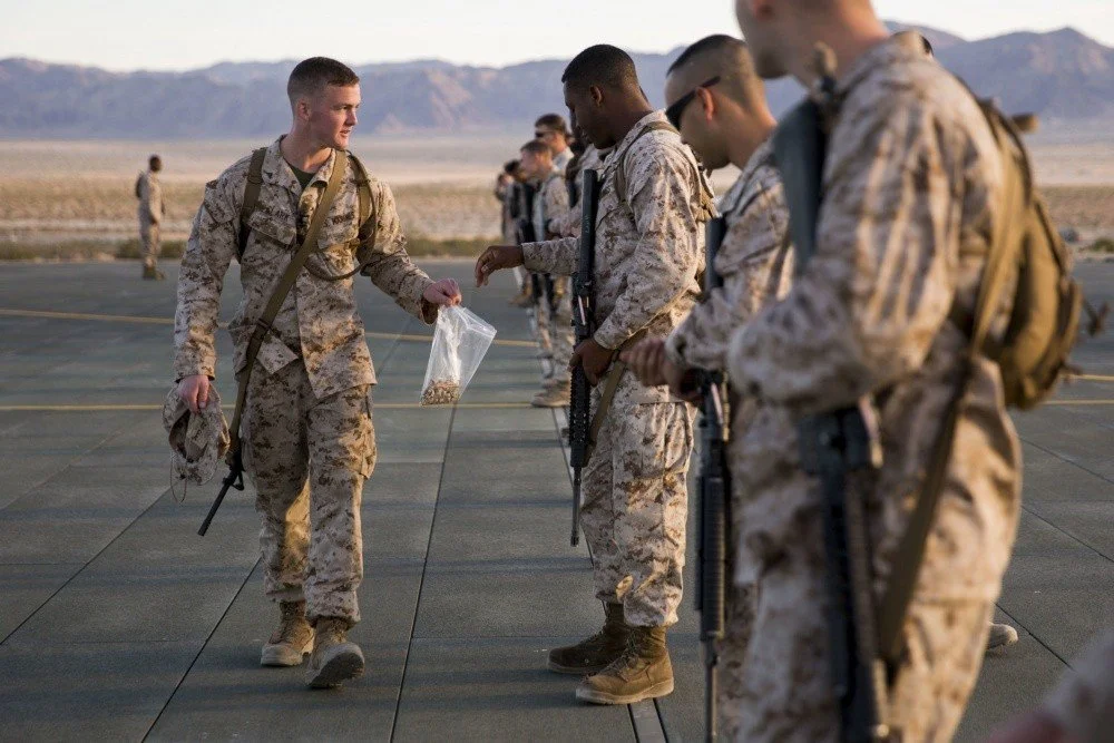 Military personnel in desert camouflage uniforms standing in line on an airstrip, one handing a plastic bag with snacks to another.