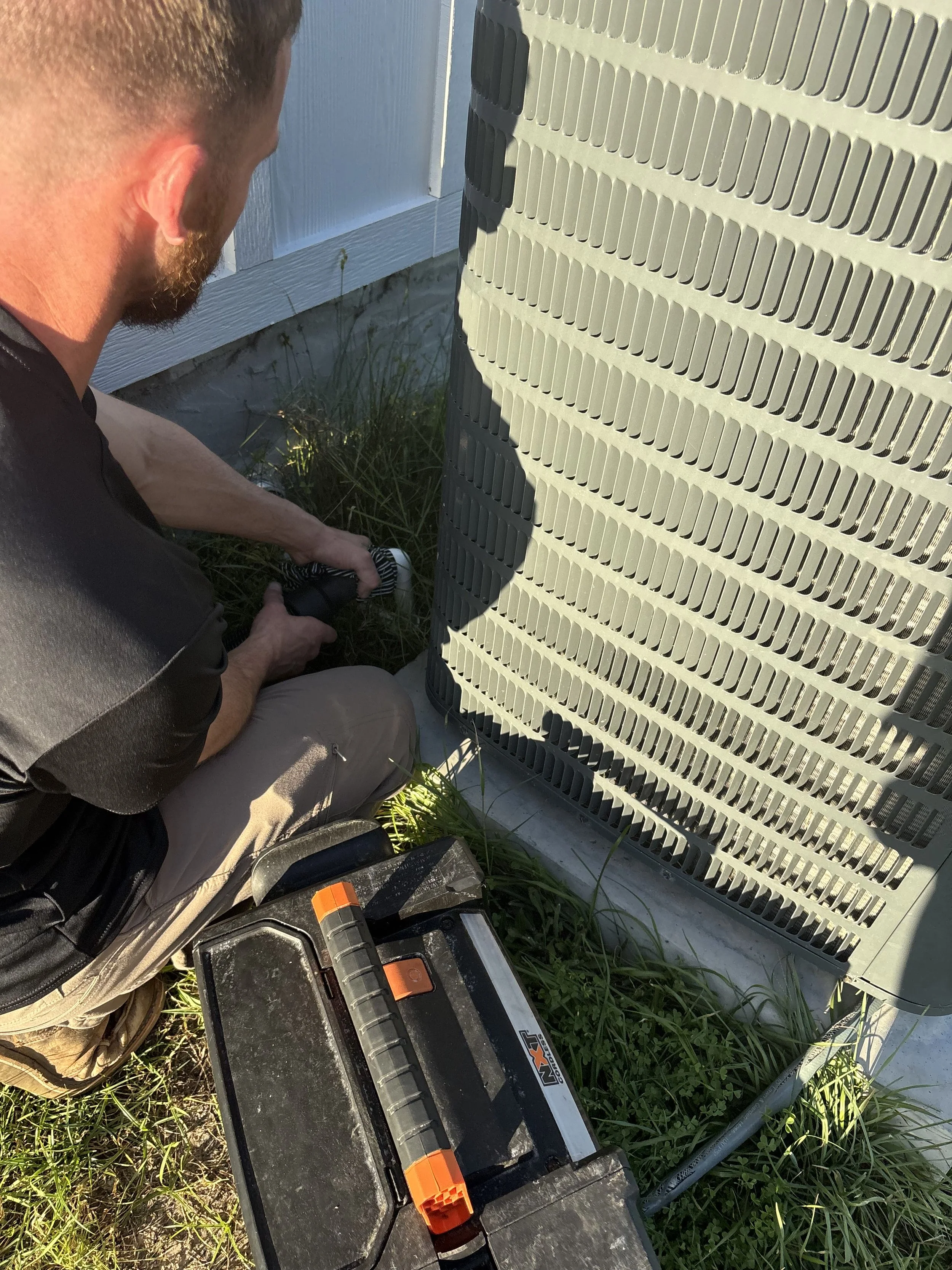 A technician working on an outdoor air conditioning unit, using tools and gauges, with a toolbox on the ground nearby.