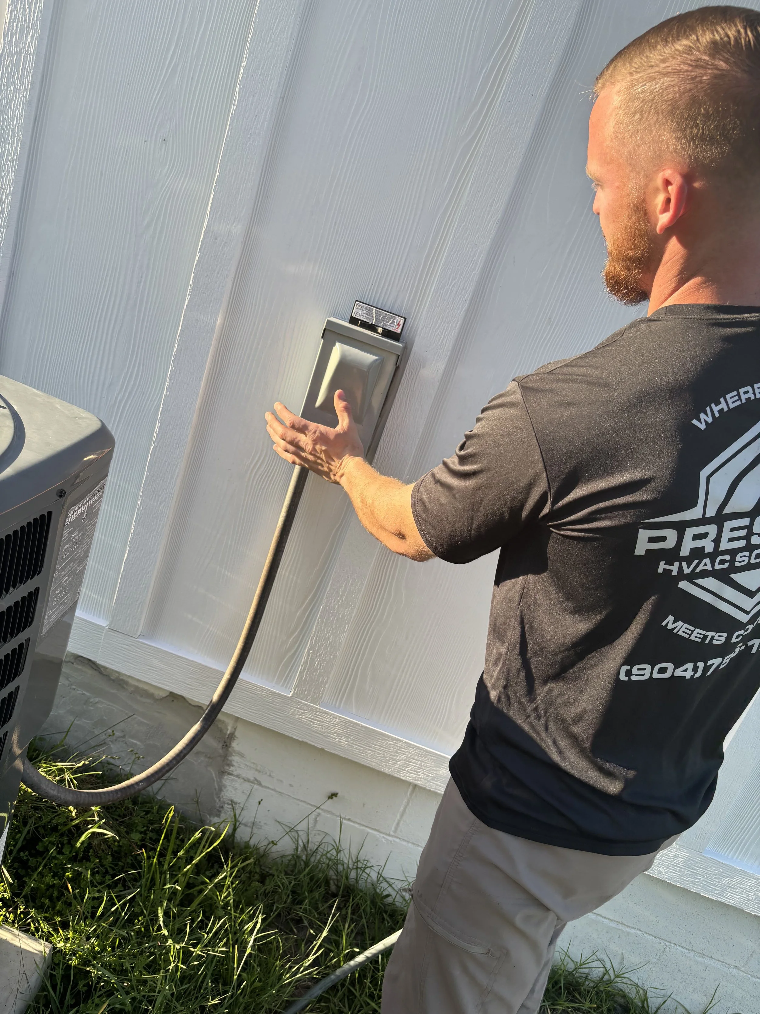 Outside HVAC unit with gray metal casing, circular fan on top, installed on a concrete pad beside a white metal building with vinyl siding, surrounded by grass and dirt.