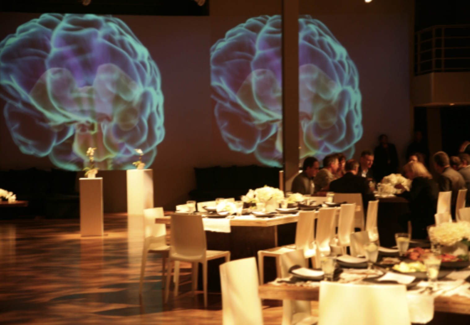 A dimly lit banquet hall with a large projection of a colorful, abstract depiction of a human brain on the wall, surrounded by guests seated at decorated tables with white chairs and floral centerpieces.