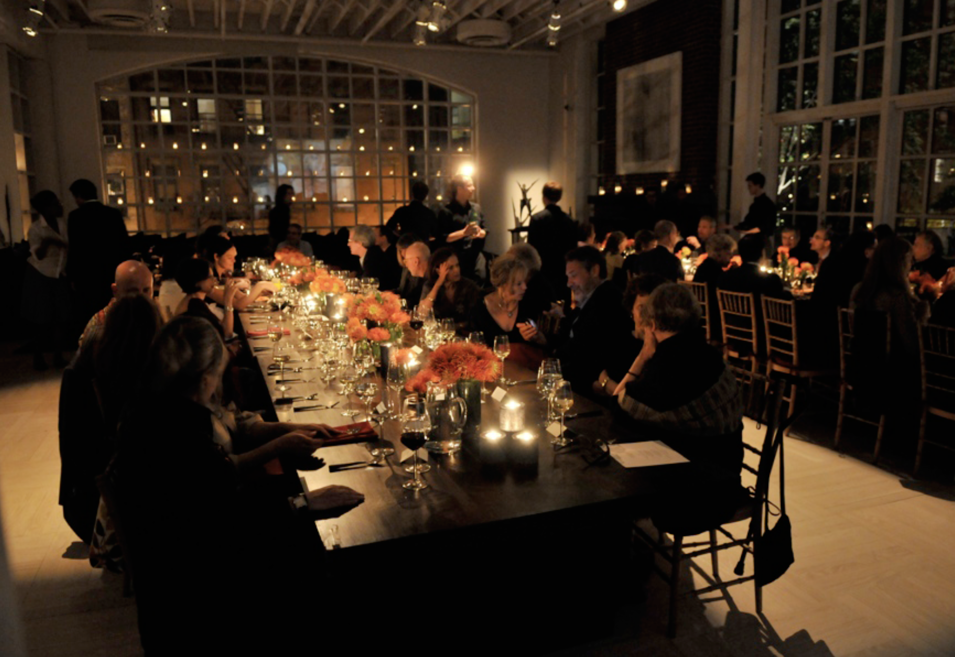 People sitting at a long dinner table in a dimly lit elegant restaurant or event space, with floral centerpieces and candlelight.