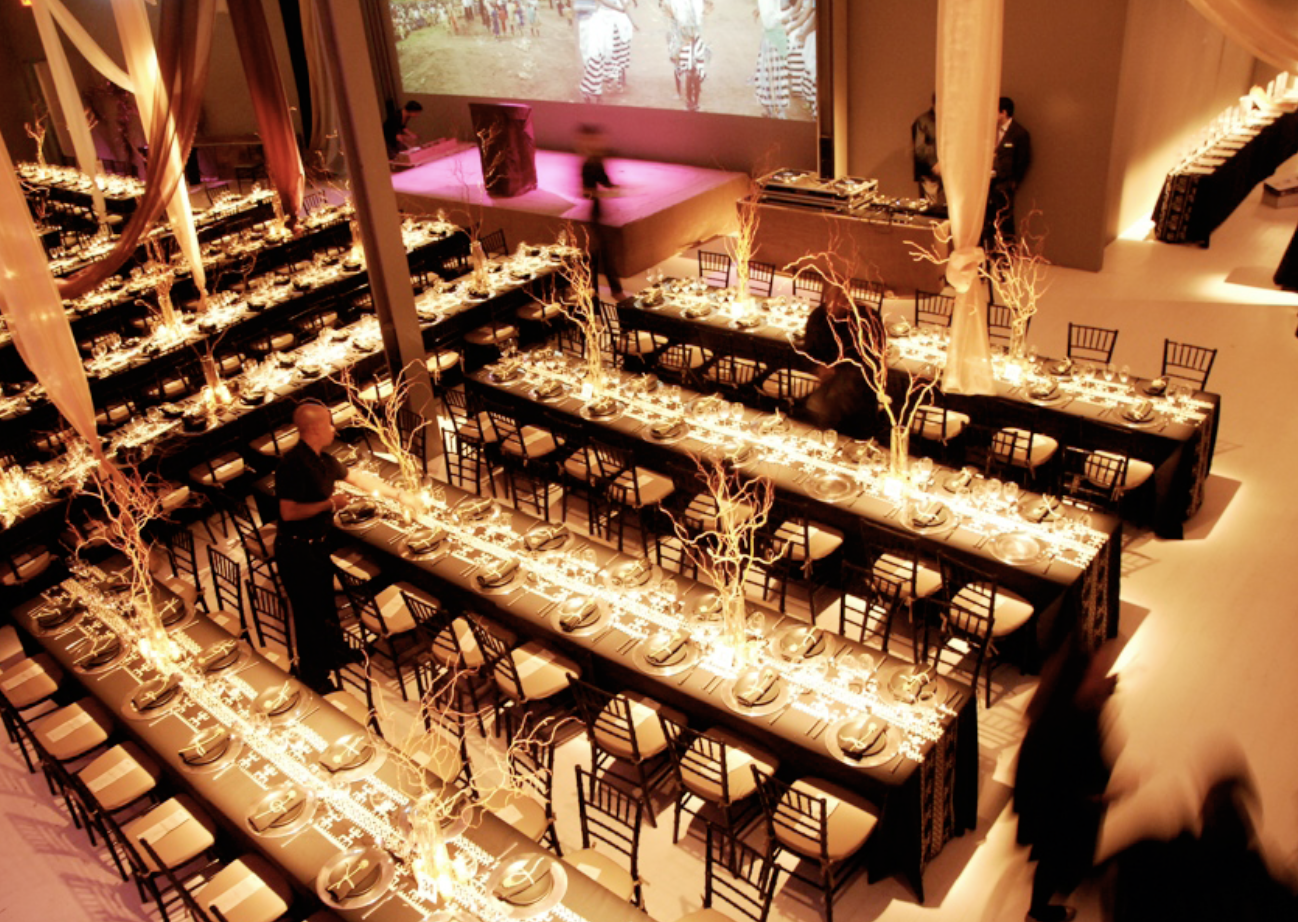 Elegant banquet hall decorated for event with long tables, beige chairs, decorative branches with lights, and a large screen displaying an outdoor scene with children.