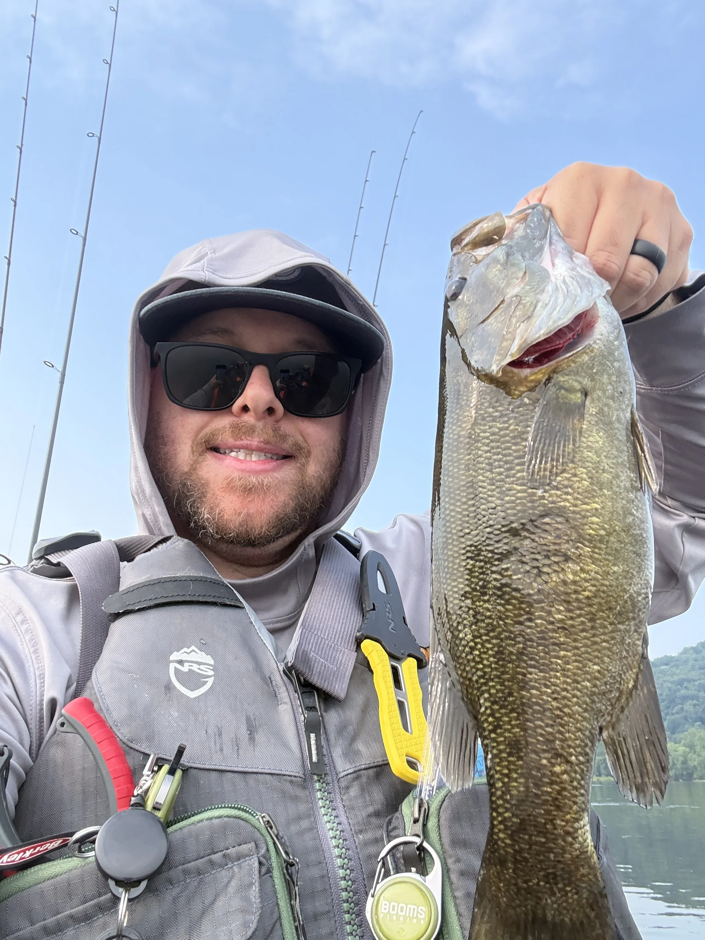 A man wearing sunglasses and fishing gear holding a large fish on a boat with fishing poles in the background under a blue sky.