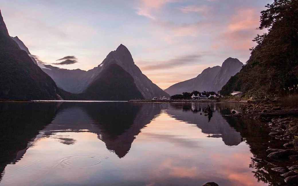 A serene landscape of Milford Sound, featuring towering mountains reflected in calm water during sunset with pink and purple sky.