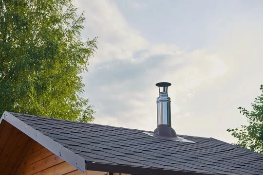 Close-up of a roof with a chimney on a house, with trees and a partly cloudy sky in the background.