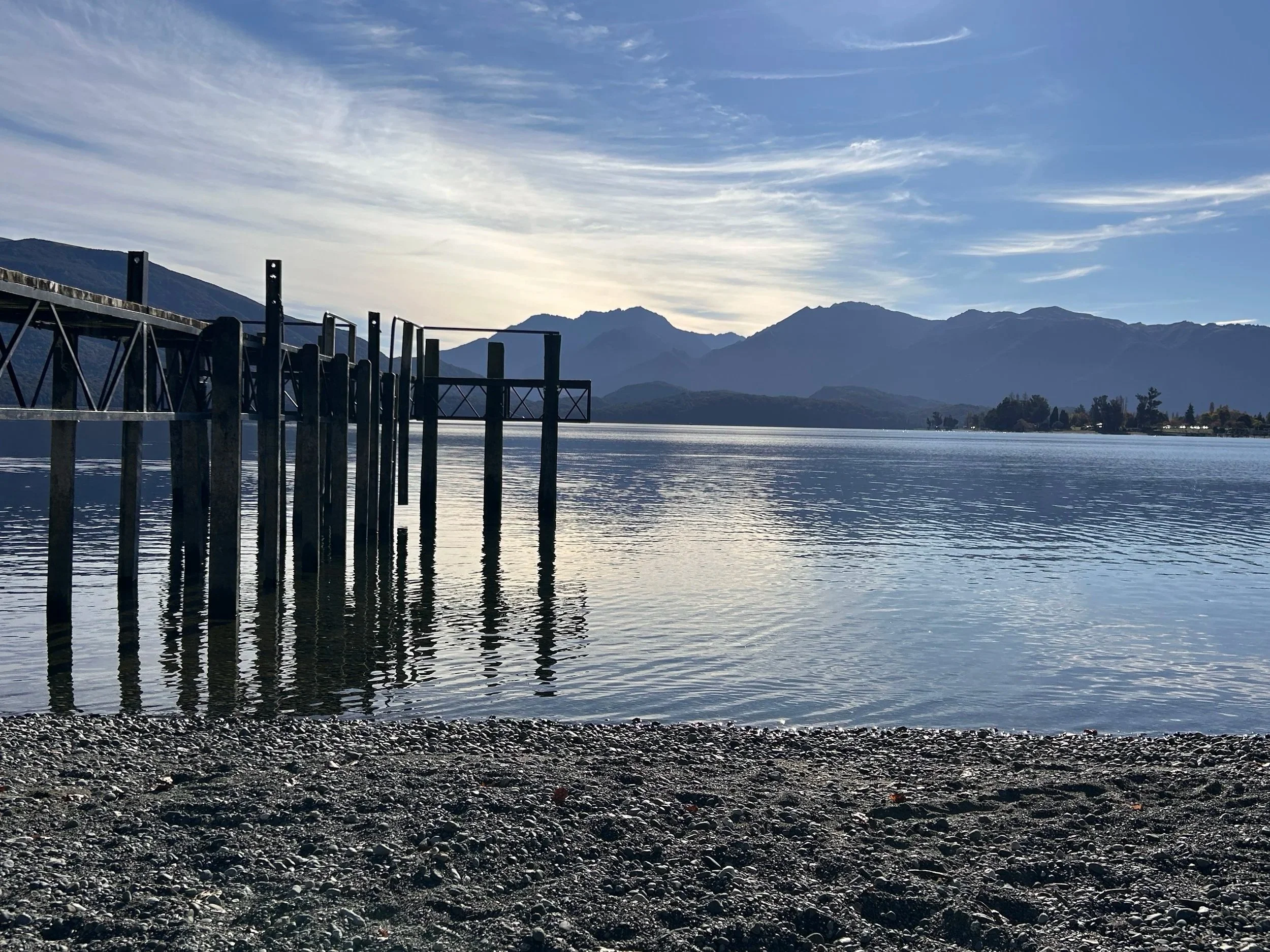 A calm lake with a wooden pier extending into the water, mountains in the background, and a partly cloudy sky at either sunrise or sunset.
