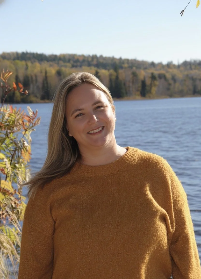 A woman with long blonde hair smiling outdoors near a lake with trees in the background.