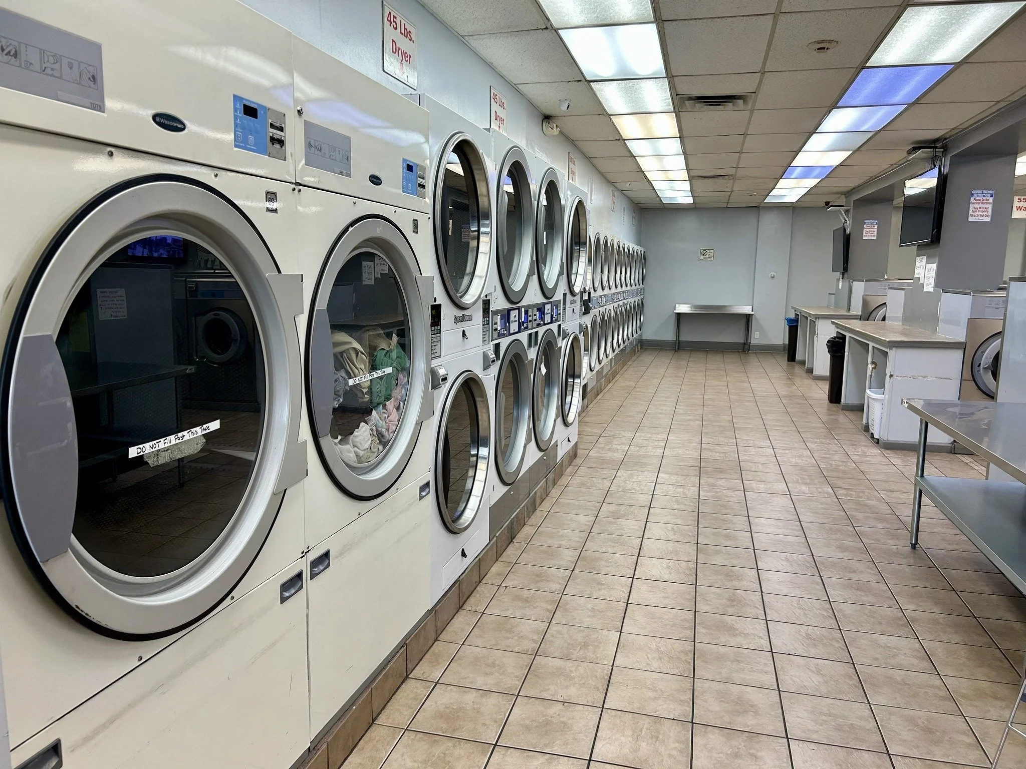 A laundromat with multiple front-loading washing machines along the left wall, some filled with clothes, and a row of dryers along the back wall. The floor is tiled, and the ceiling has fluorescent lighting. There are no people in the image.