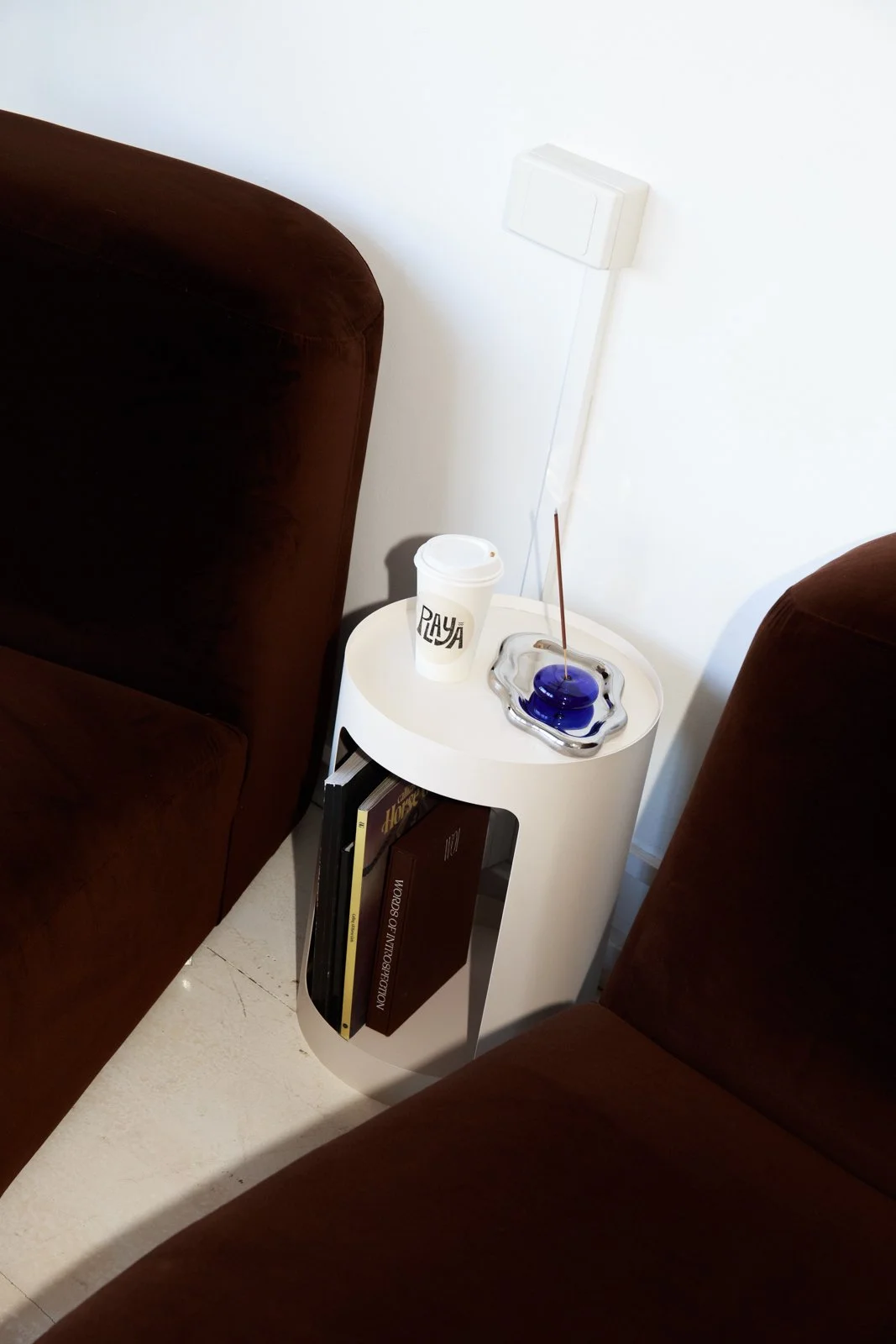 A white side table with a white travel mug, a blue glass ashtray with a cigarette, and a brown book. The table is placed between two brown upholstered chairs or sofas against a white wall with a power outlet.