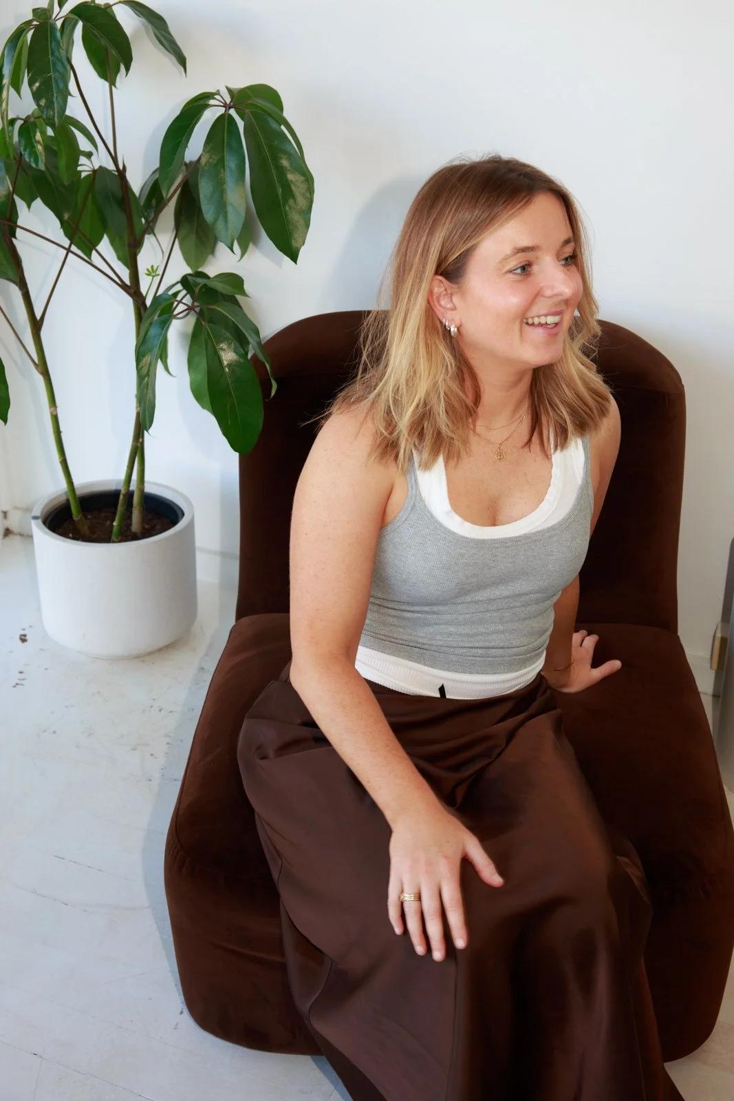 A woman with blonde hair and earrings sitting on a brown chair, smiling, with a large potted plant next to her in a white pot, against a white wall.