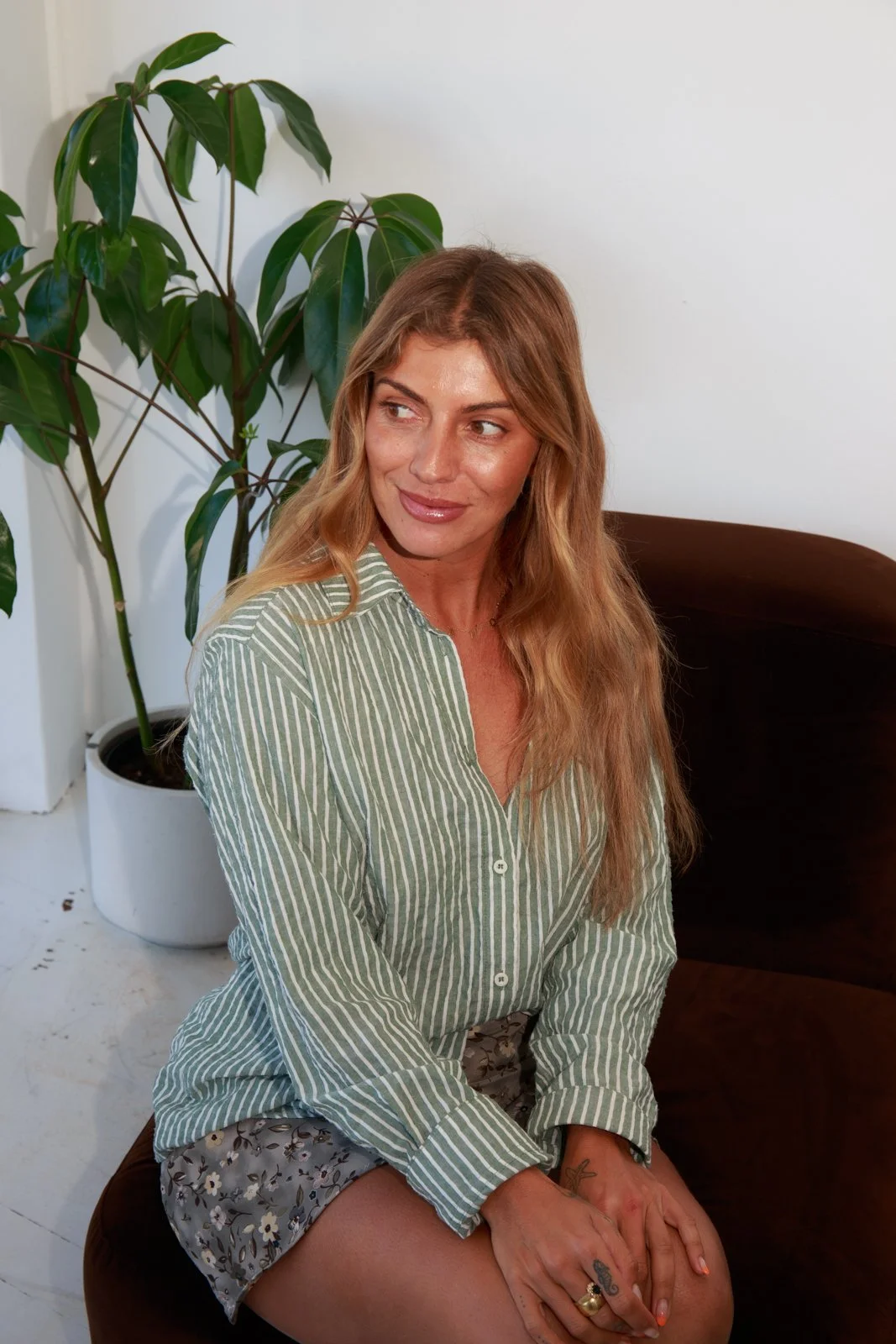 A woman with long wavy blonde hair, wearing a green and white striped shirt and floral shorts, sitting on a brown chair in front of a large potted plant in a room with white walls.