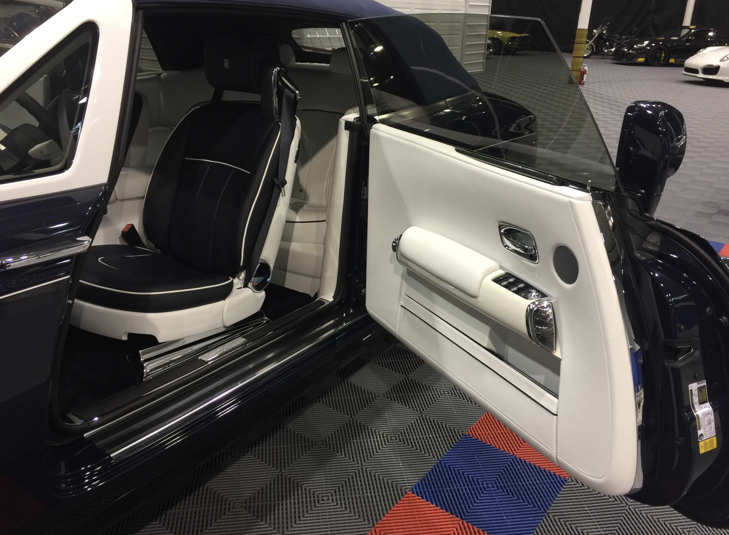Interior of a luxury car showing a black and white leather seat, white door panel with chrome accents, controls, and a speaker, with other parked cars visible outside in a garage or showroom setting.