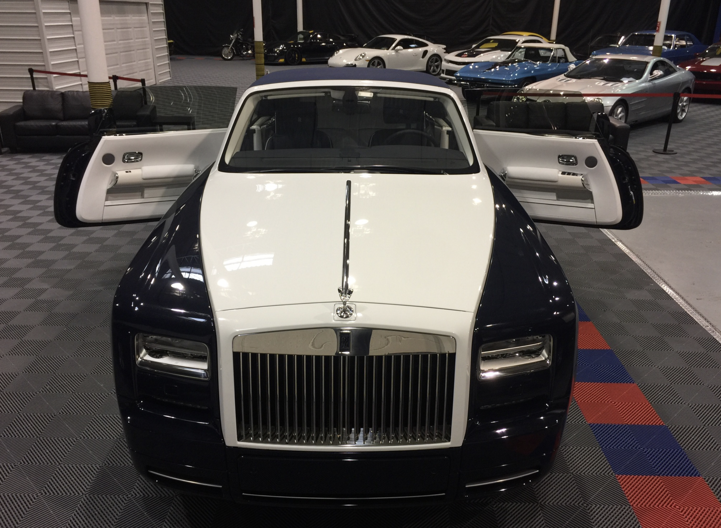 A black and white Rolls Royce car with open doors inside a showroom filled with luxury cars.