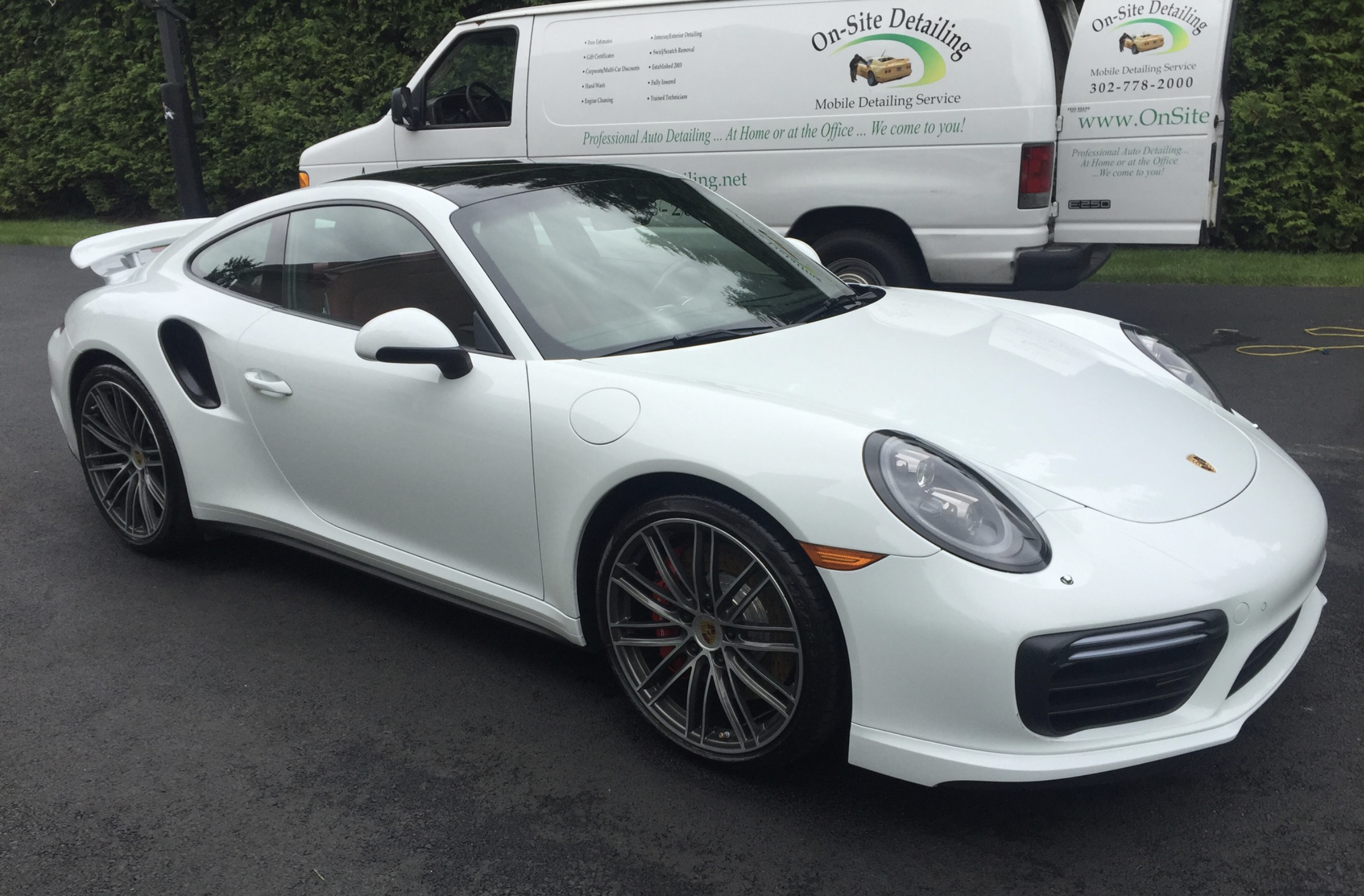 A white Porsche 911 sports car parked on a black asphalt surface, with black multi-spoke wheels, behind a white van advertising on-site detailing service.