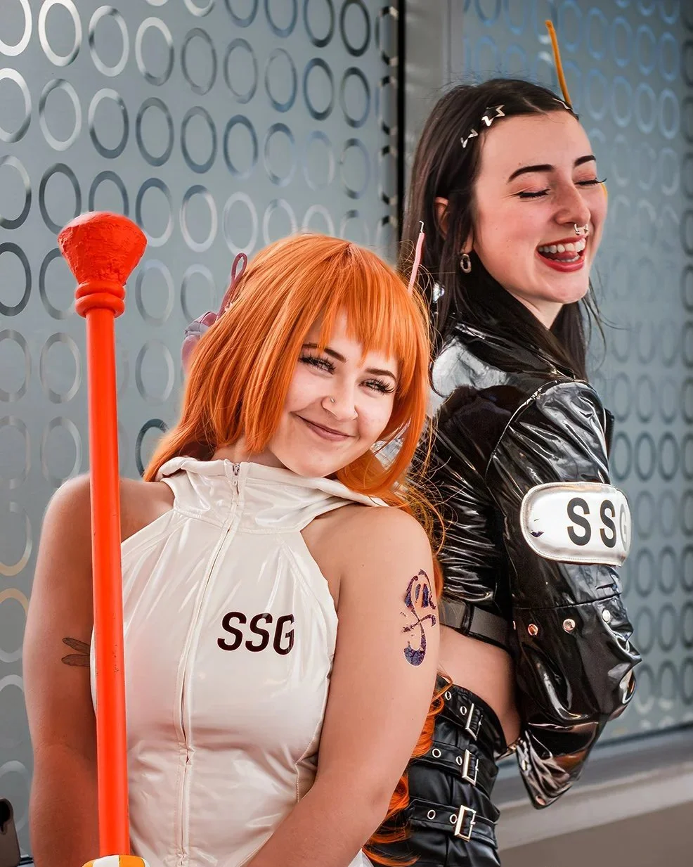 Two young women dressed in cosplay costumes with 'SSG' patches, smiling and leaning against a metallic wall with circular patterns.
