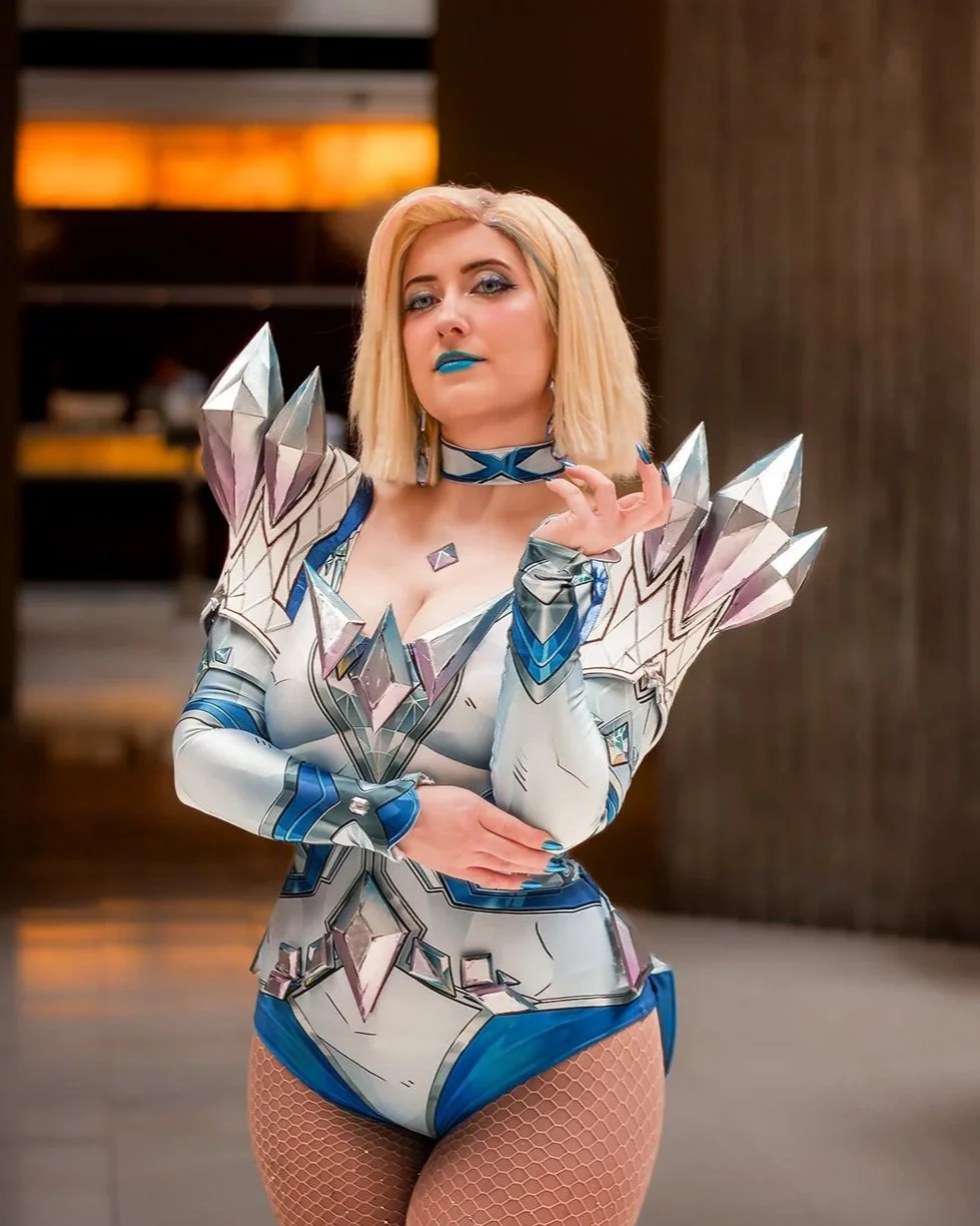 Woman in a futuristic, armored costume with reflective, crystal-like shoulder accents, blue lipstick, fishnet stockings, and blonde bob haircut.