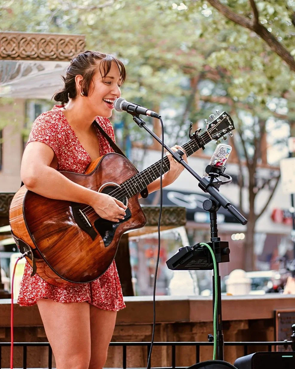 A woman with brown hair in a bun, wearing a red floral dress, playing an acoustic guitar and singing into a microphone outdoors. A smartphone on a stand is recording her performance.