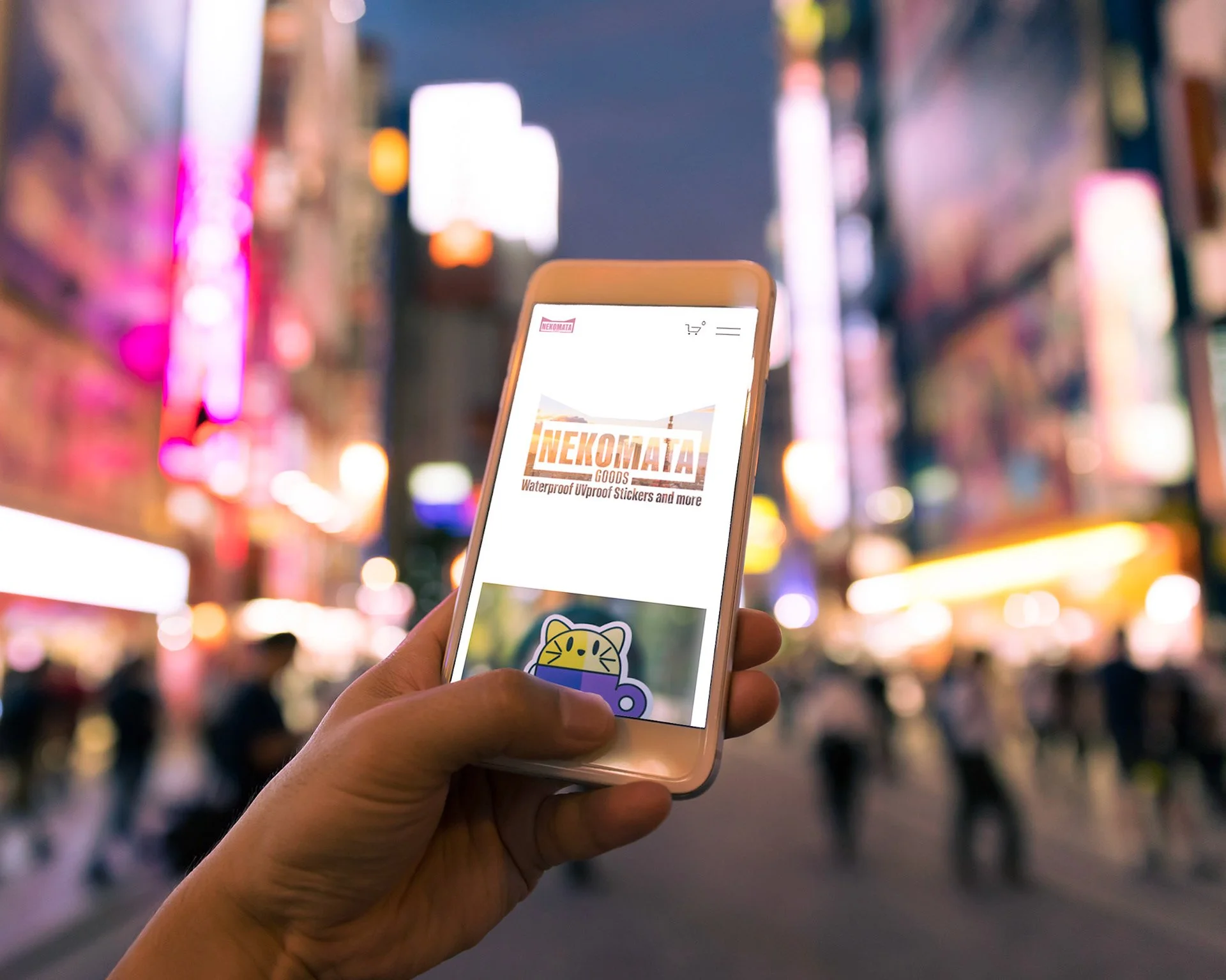 Person holding smartphone displaying Nekomata Goods website in a busy city street at night with colorful neon lights and blurred crowd.