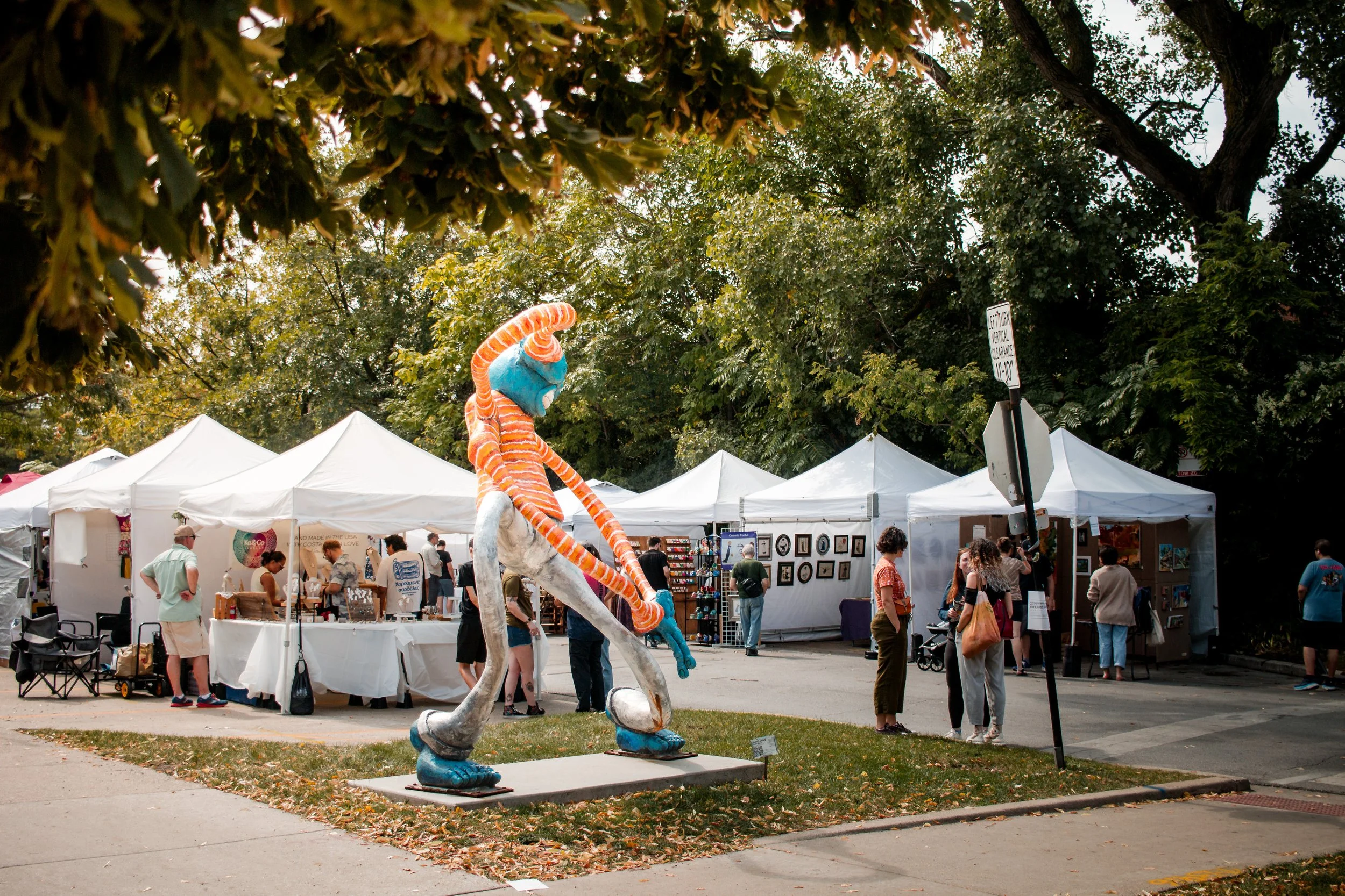 An outdoor art fair with white tents, people browsing booths, and a tall, colorful sculpture of a whimsical character with striped orange and blue features, set against a backdrop of trees.