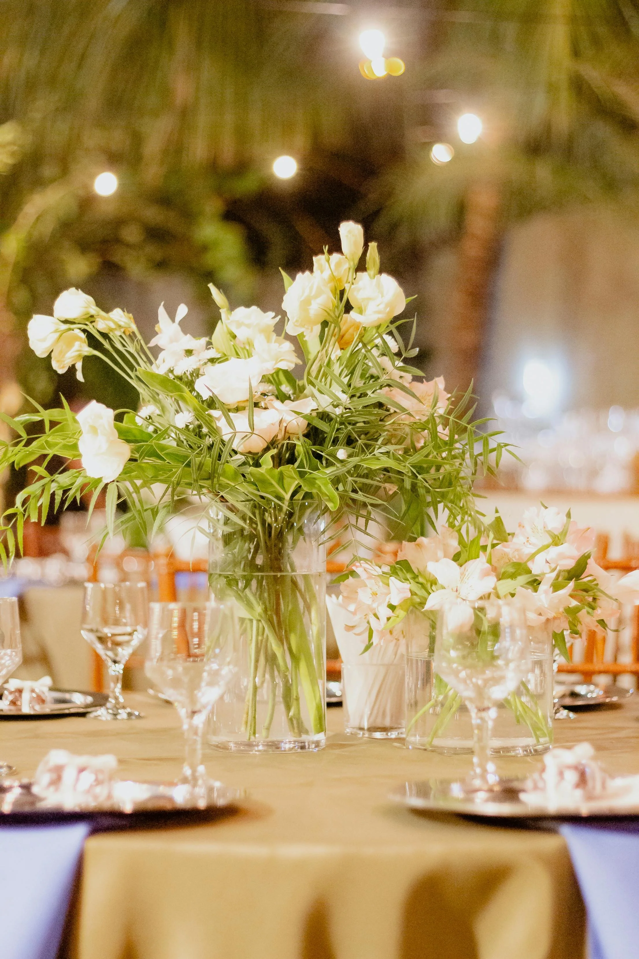 A table centerpiece with white and pale pink flowers in clear glass vases, surrounded by wine glasses and place settings, in an indoor dining setting.