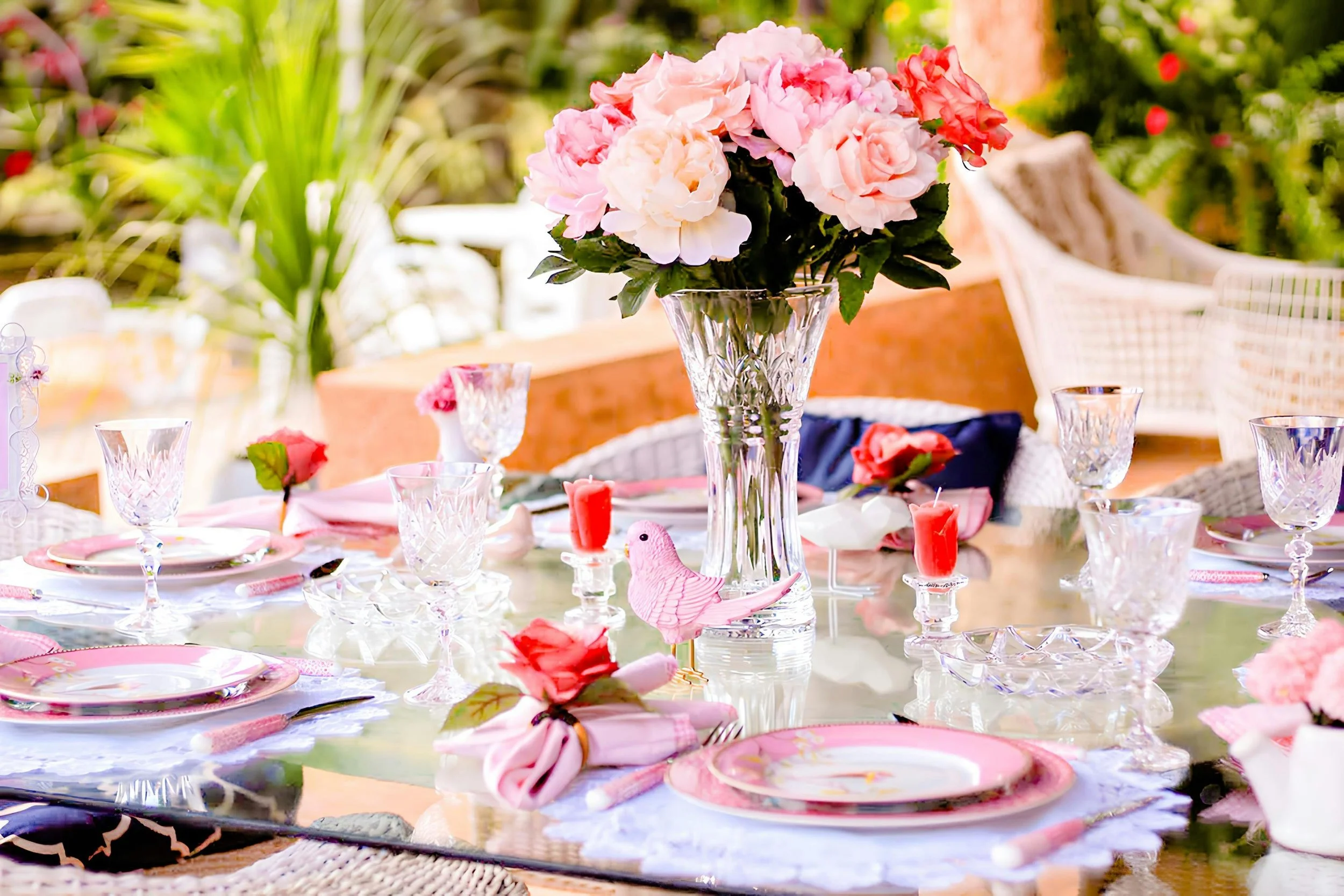 A decorated outdoor dining table with a large pink and white floral arrangement in a tall glass vase, crystal glassware, pink and red candles, pink plates, and pink napkins, surrounded by white chairs and lush green plants in the background.
