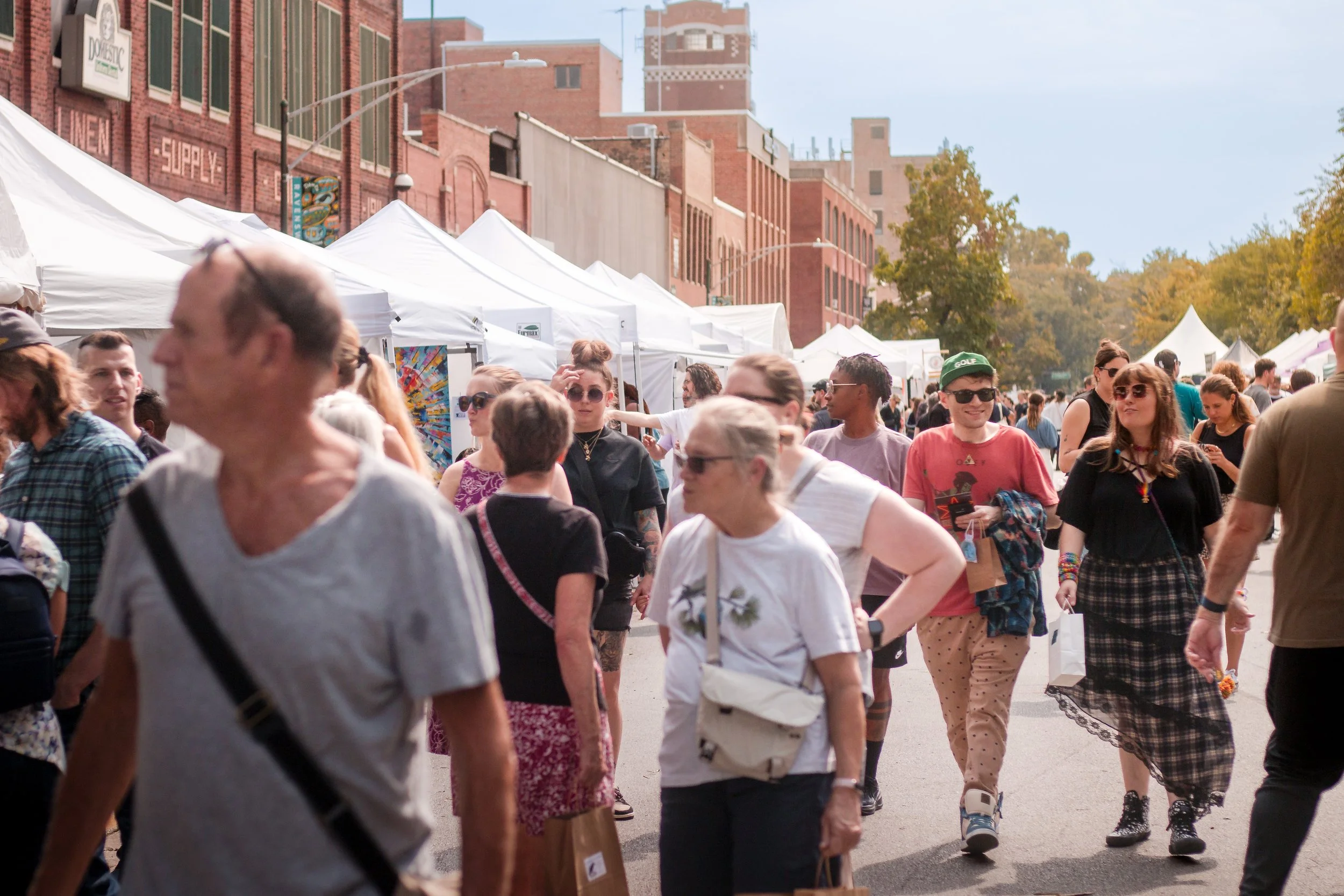People walking among vendor tents at an outdoor street market during daytime with brick buildings and trees in the background.