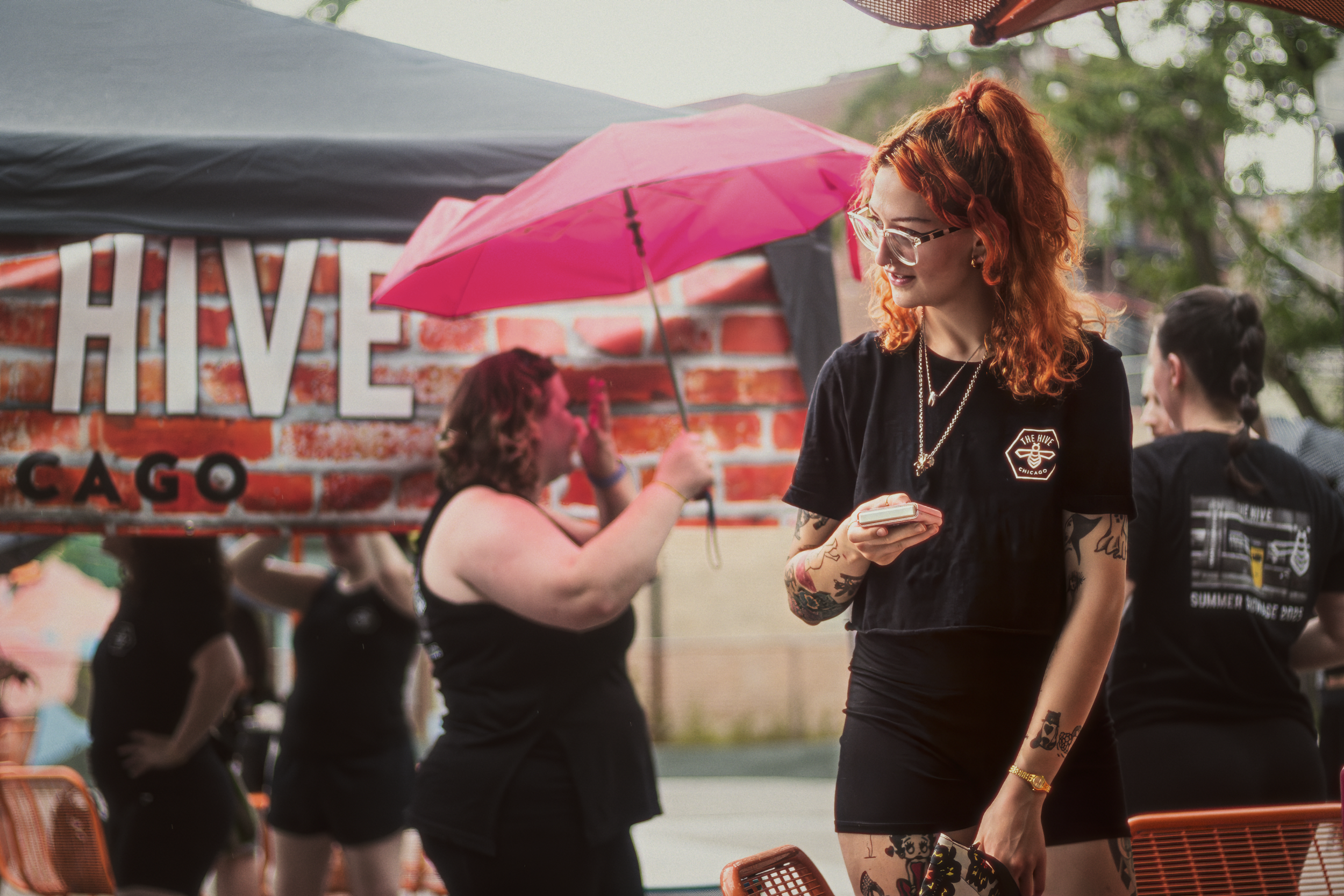 A woman with red hair and tattoos standing outdoors at a festival, looking at her phone, with other people and a brick wall with a sign in the background.