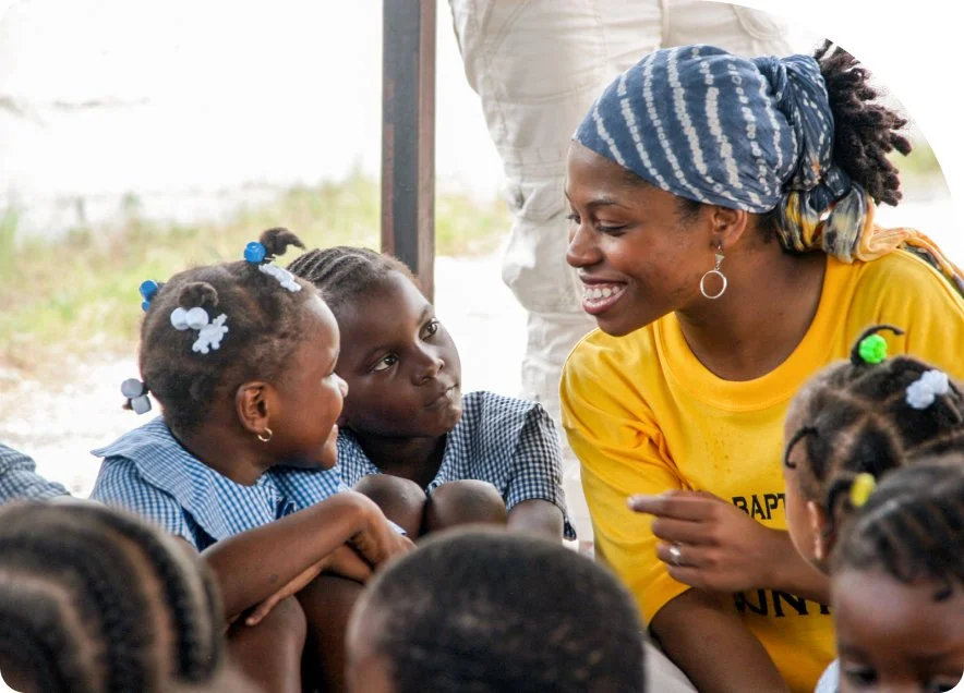 A woman in a yellow shirt and blue headscarf interacts with young children in a classroom setting, with outdoor scenery in the background.