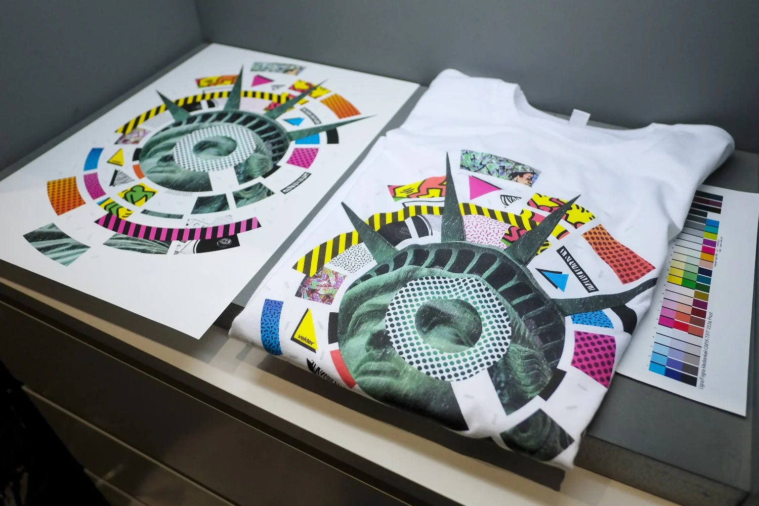 Colorful graphic design artwork of the Statue of Liberty's face and crown, printed on paper and T-shirt, displayed on a desk with color calibration charts.