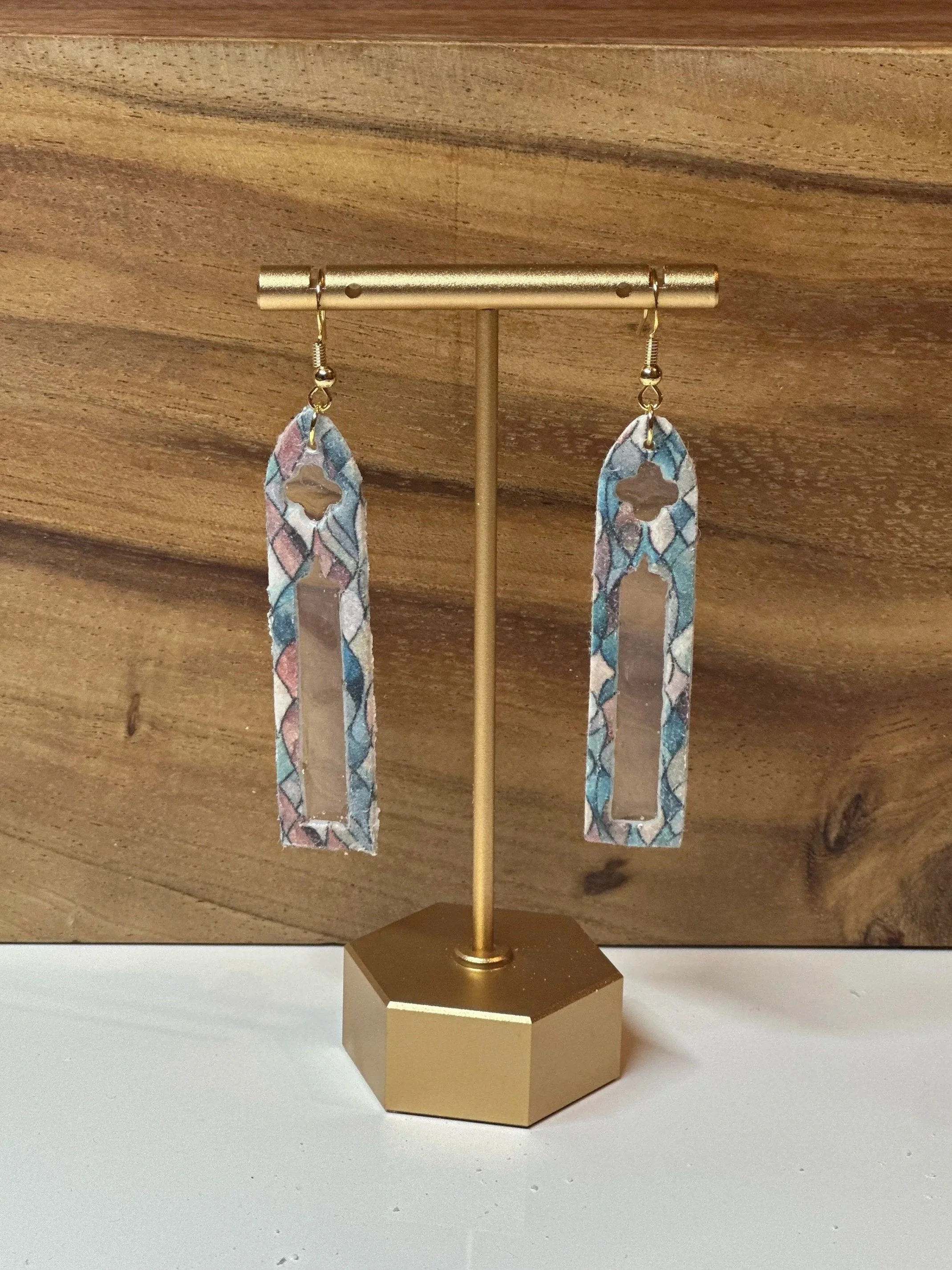 Pair of colorful, patterned, rectangular earrings with geometric cutouts, hanging on a gold display stand.