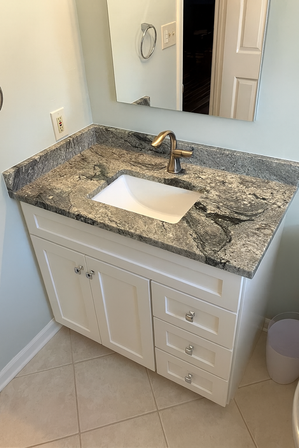 Bathroom vanity with a granite countertop, a white sink, metallic faucet, mirrored wall, electrical outlet, and white cabinetry, with a glimpse of a closet door in the background.