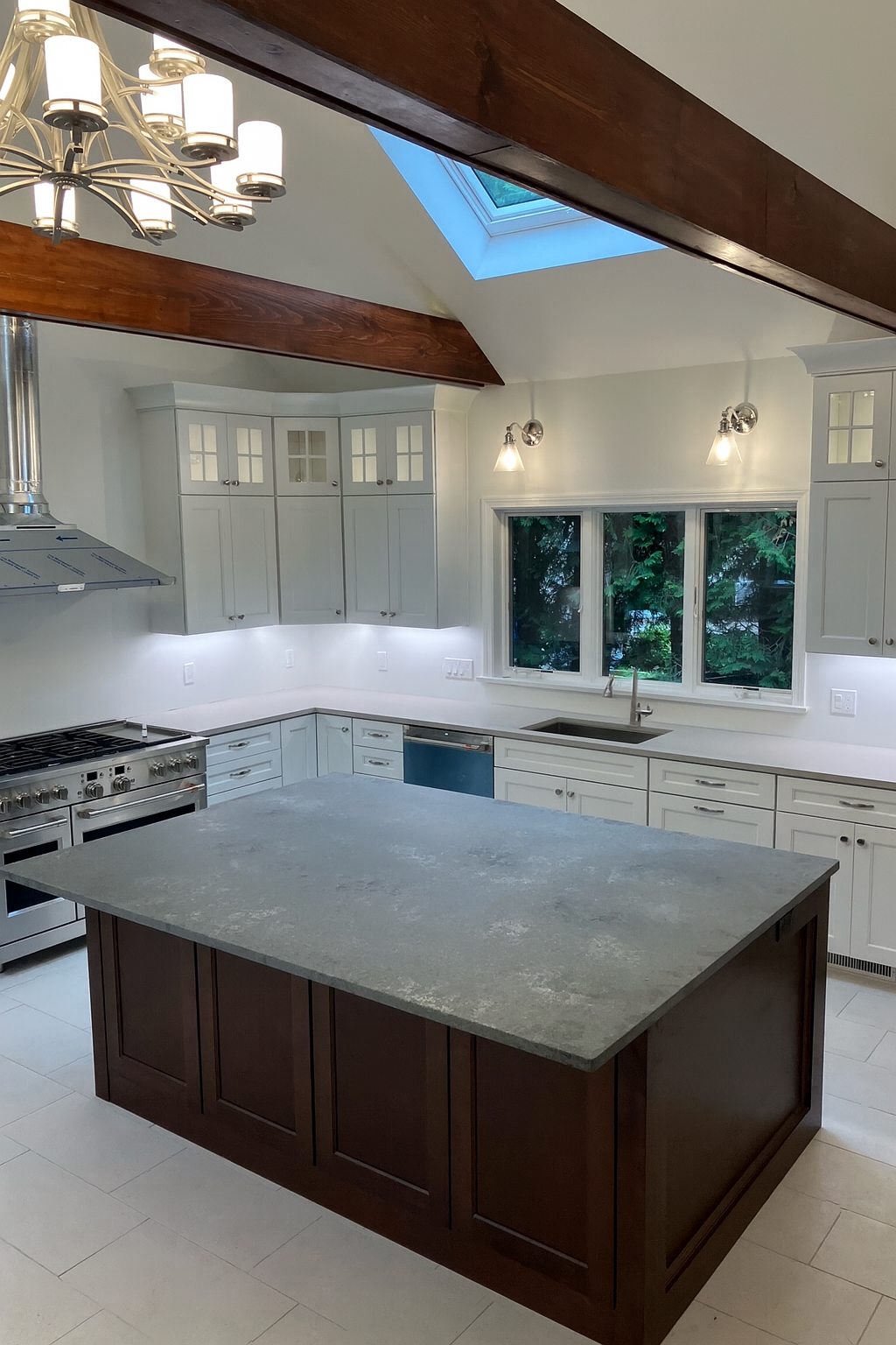 Modern kitchen with white cabinetry, a large gray island with wooden base, stainless steel stove, and a skylight in a vaulted ceiling with wooden beams.