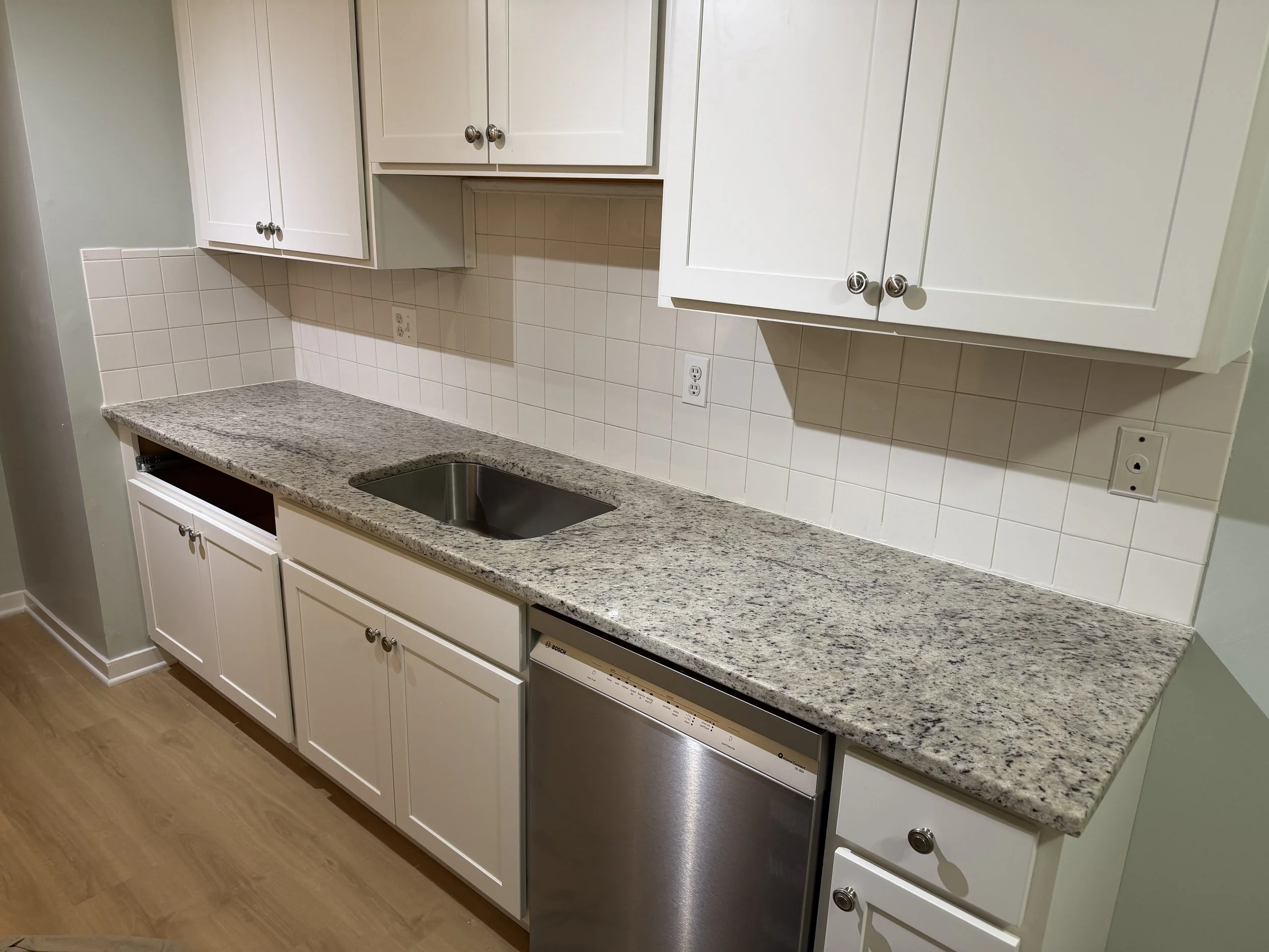 A kitchen countertop with white cabinets, a stainless steel sink, a dishwasher, tiled backsplash, and electrical outlets.