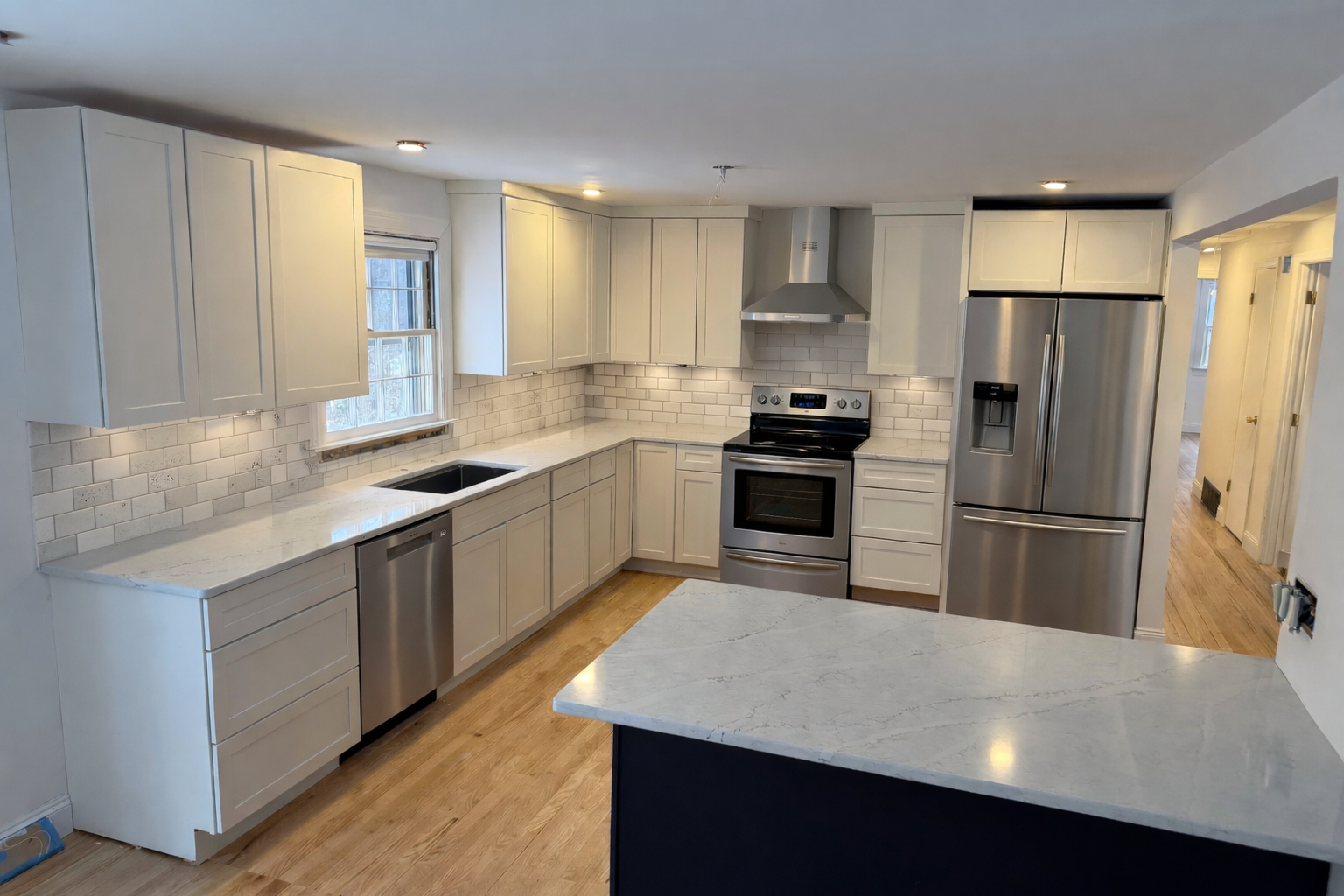 Modern kitchen with white cabinets, stainless steel appliances, a window above the sink, light-colored marble countertops, wood flooring, and white subway tile backsplash.