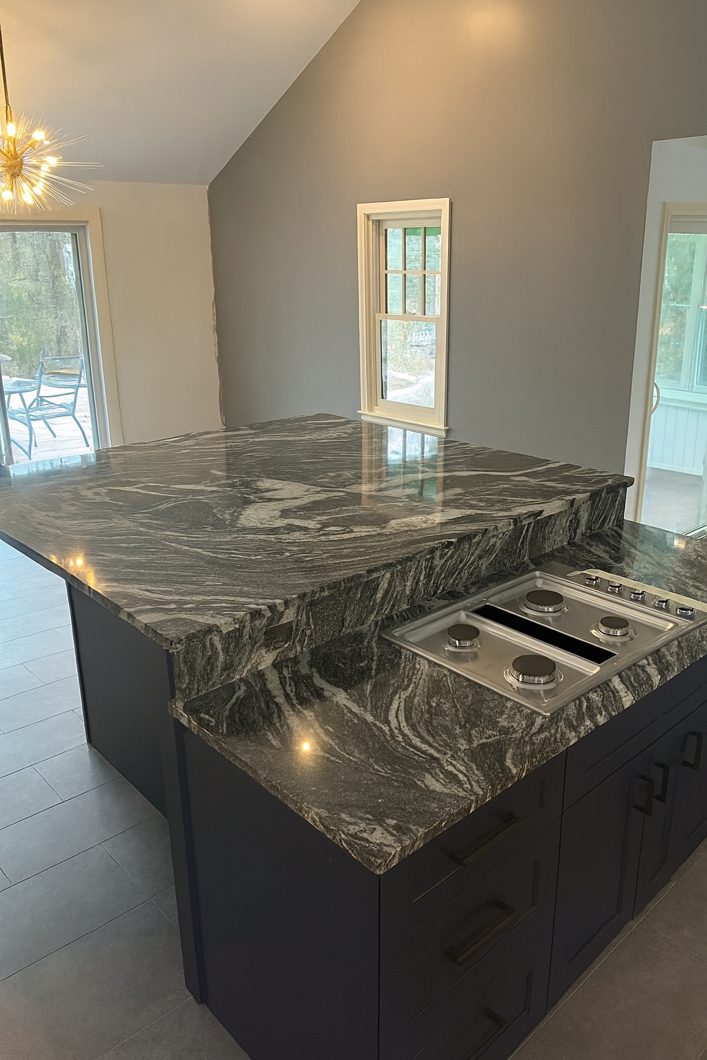 Kitchen with dark granite countertop on island, stove with four burners, light-colored walls and windows, sliding door leading to outdoor area, and a light fixture hanging from the ceiling.
