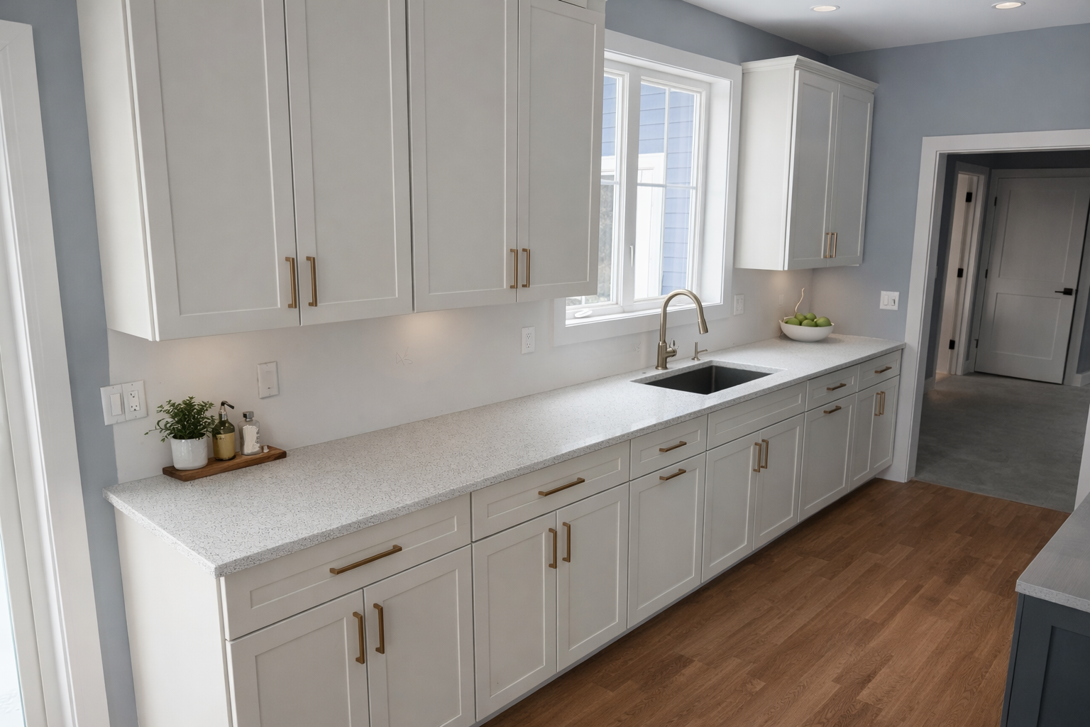 Modern kitchen with white cabinets, a white speckled countertop, a window above the sink, a black undermount sink, and gold handles. Small potted plant, soap, and a bowl of green apples on the countertop.
