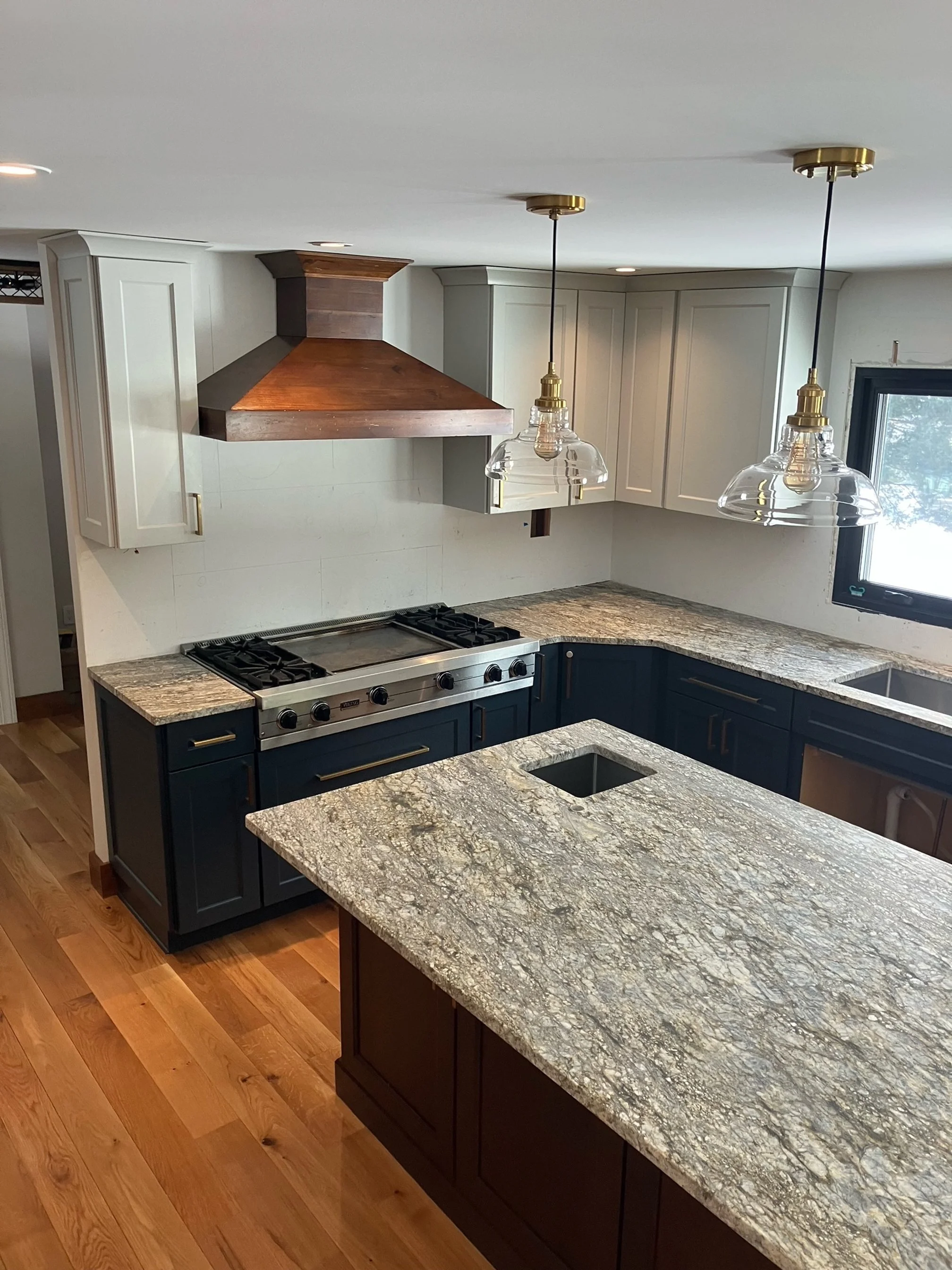 Modern kitchen with granite countertops, navy blue lower cabinets, white upper cabinets, a stainless steel stove, wooden range hood, and two glass pendant lights.