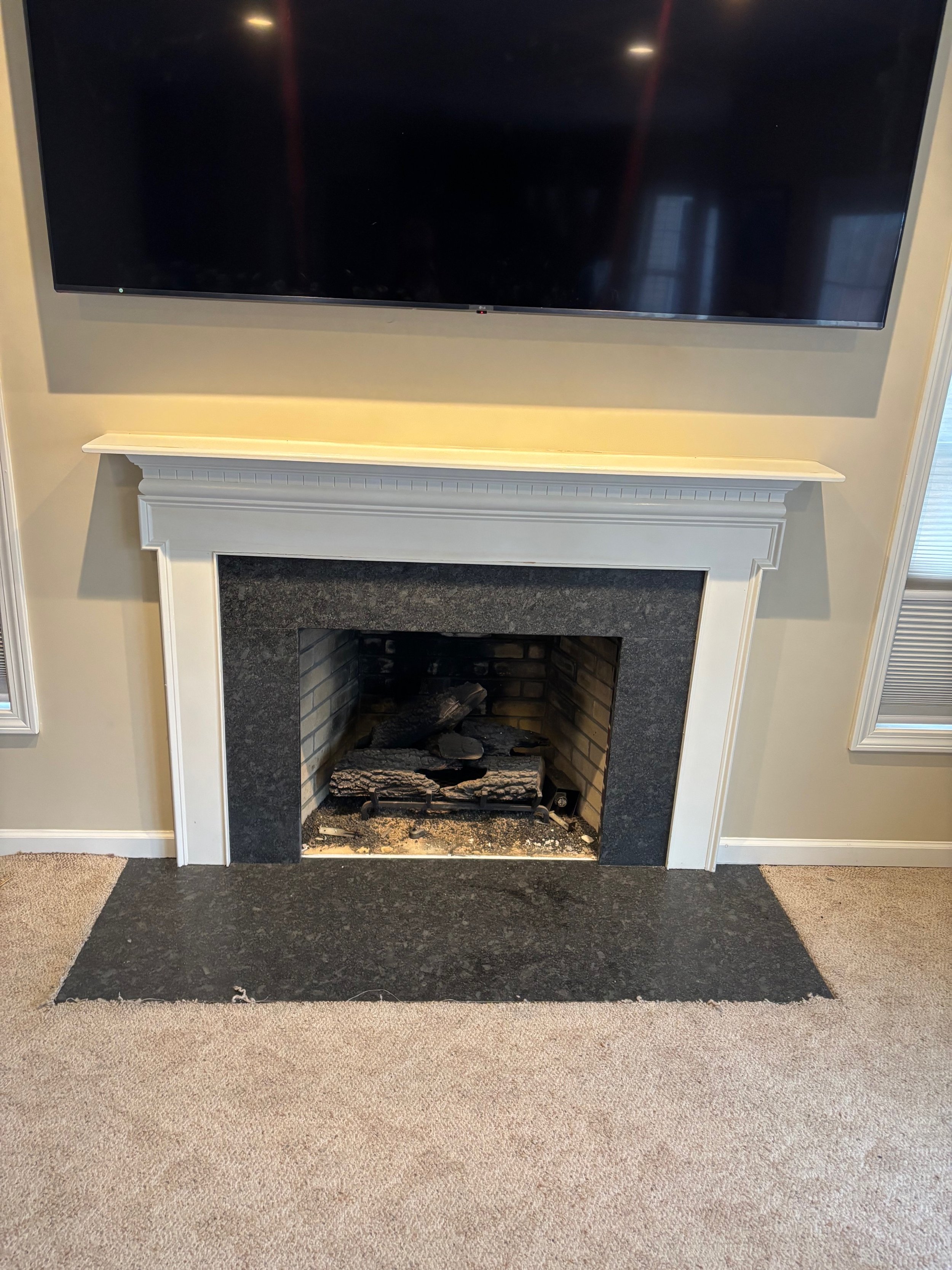 Living room fireplace with black granite surround and a mounted flat-screen TV above, beige walls, and beige carpeted floor.