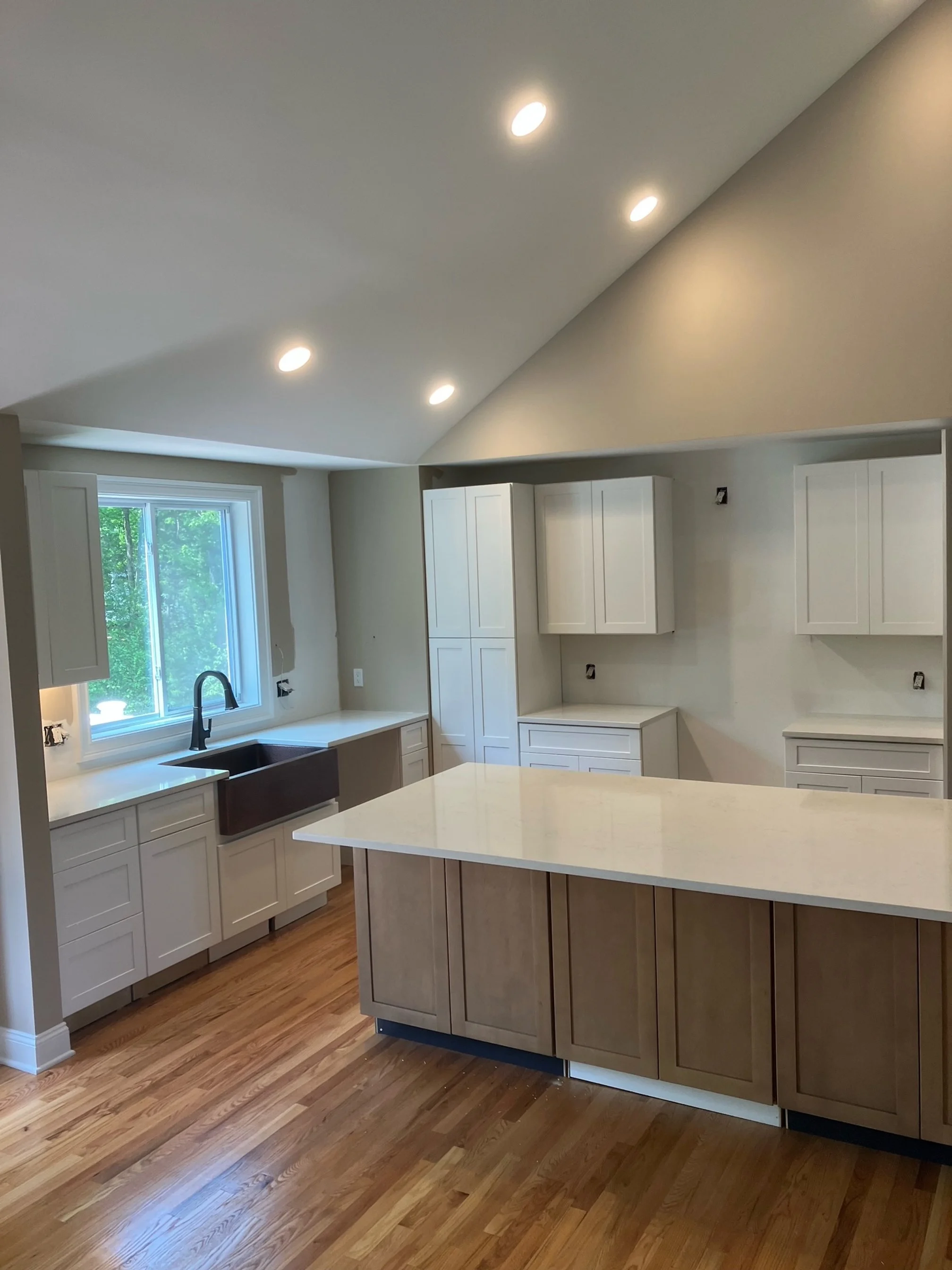 Empty kitchen with white cabinets, a central island with wooden cabinets, a black sink under a window, hardwood floors, and recessed ceiling lights.