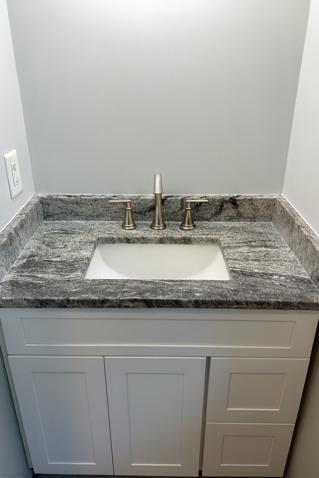 Bathroom vanity with a granite countertop, a built-in sink, and a brushed nickel faucet with two handles. White cabinet with paneled doors underneath. Plain wall behind the countertop.