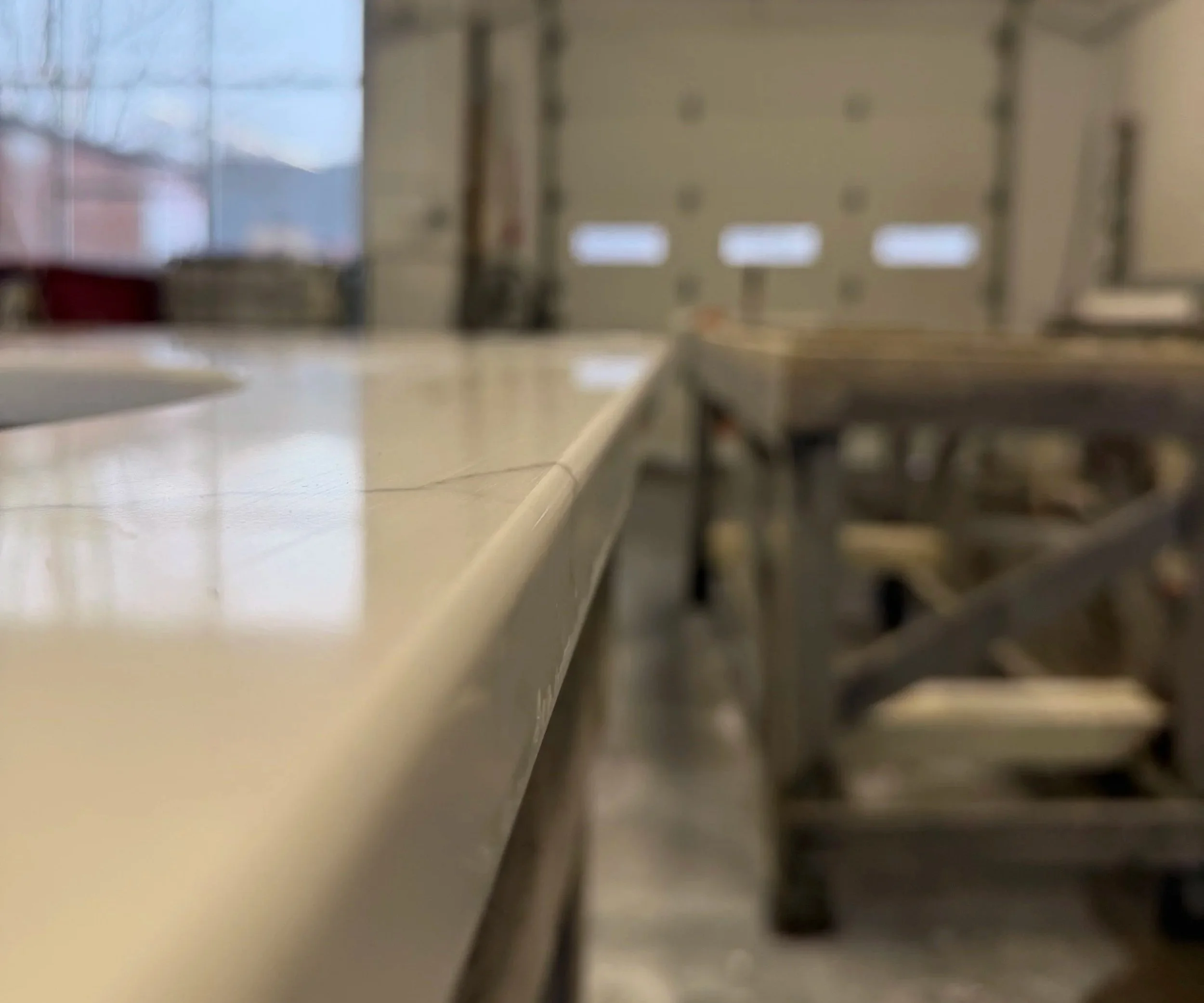 A blurry image of a workshop or garage with a large metal worktable in the foreground and a wall with windows and closed garage doors in the background.