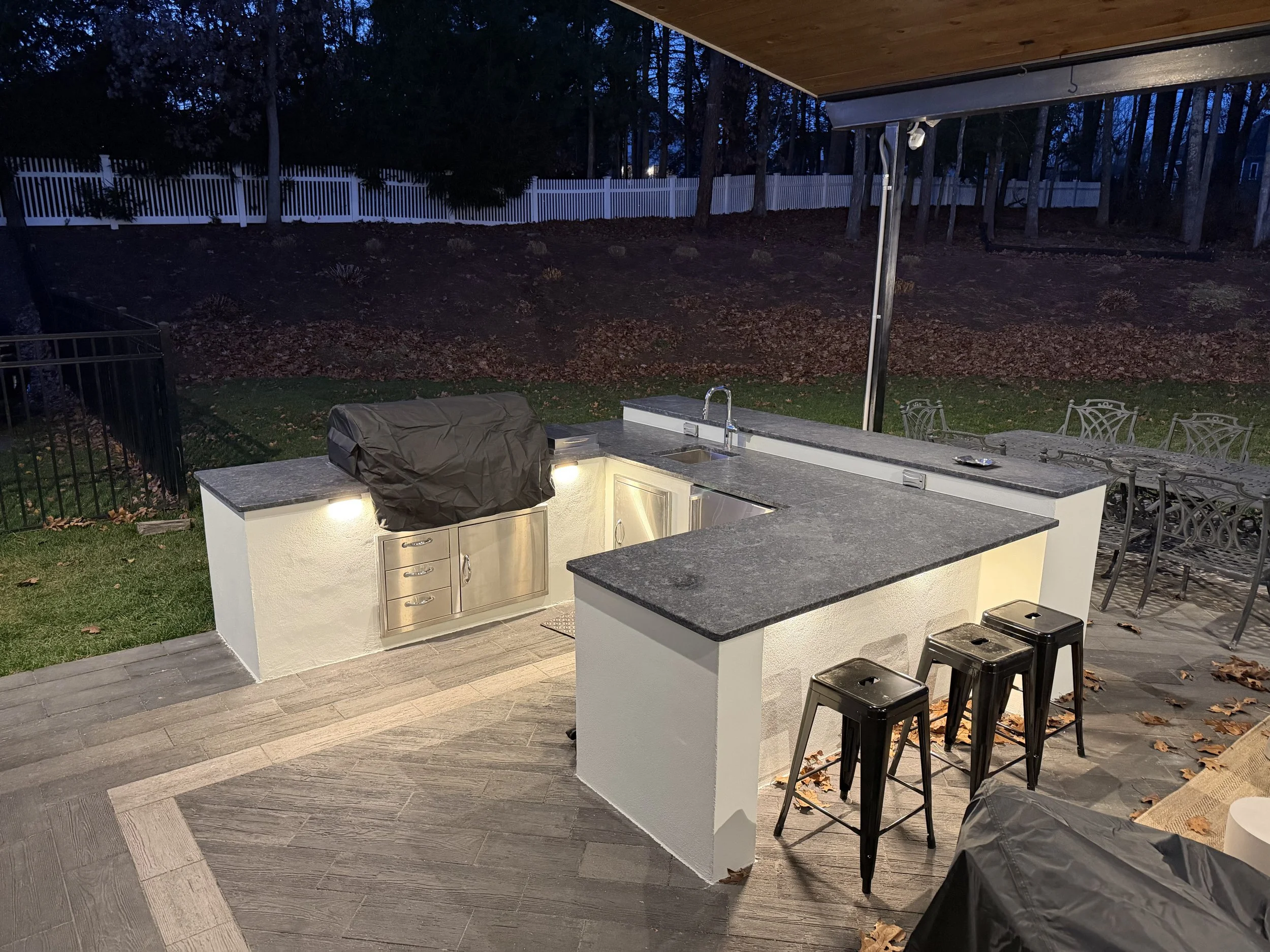 Outdoor kitchen with a grill covered by a black cover, stainless steel cabinetry, granite countertops, a sink, and three black stools. A dining table with chairs is visible in the background, with a grassy yard, trees, and a white fence beyond.