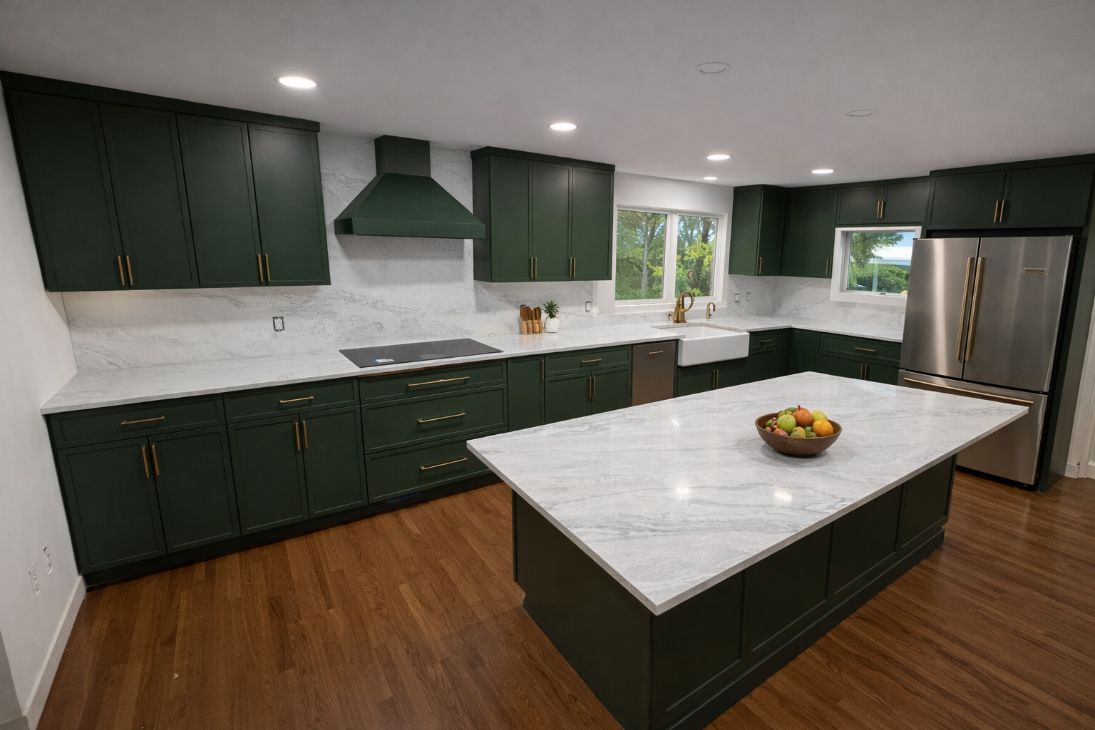 Modern kitchen with dark green cabinets, white marble countertops, a large island with a bowl of fruit, stainless steel appliances, and wood flooring, with trees visible through the windows.